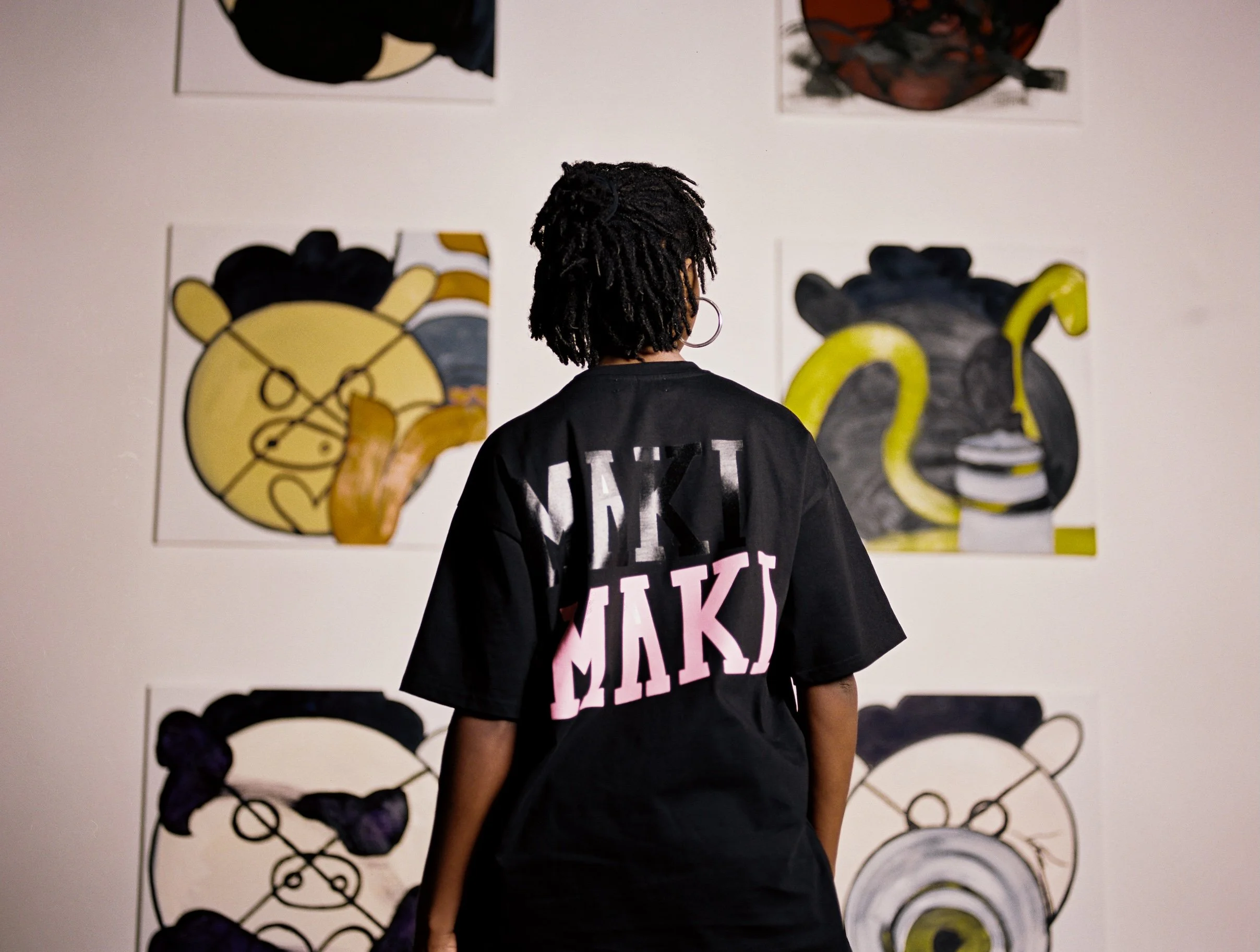 A person with short, dreadlocked hair wearing a black T-shirt with pink and white text, standing in front of a wall with colorful abstract artwork.