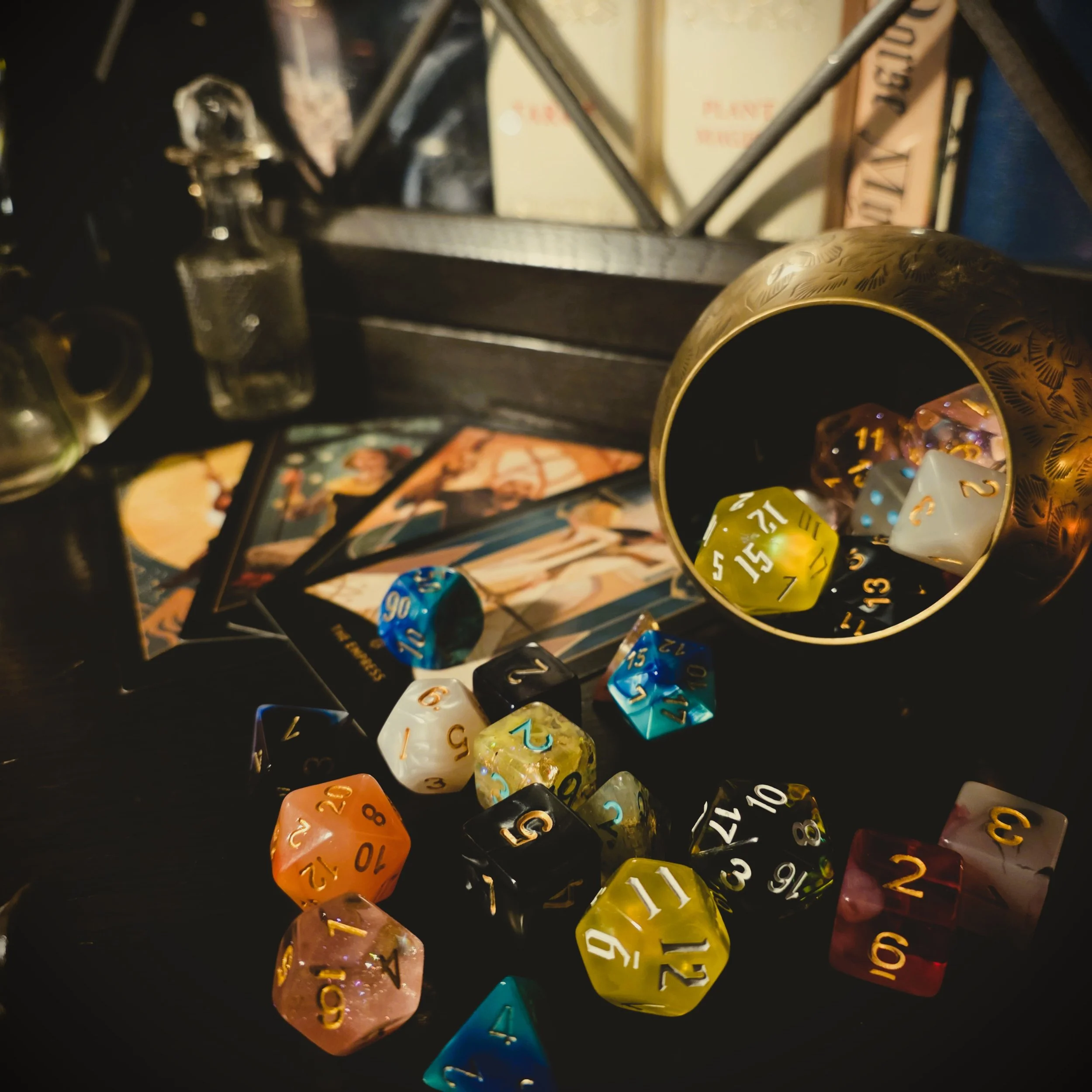 Various colorful polyhedral dice, some spilling from a metallic container, with tarot cards and vintage perfume bottles in the background, on a dark surface.