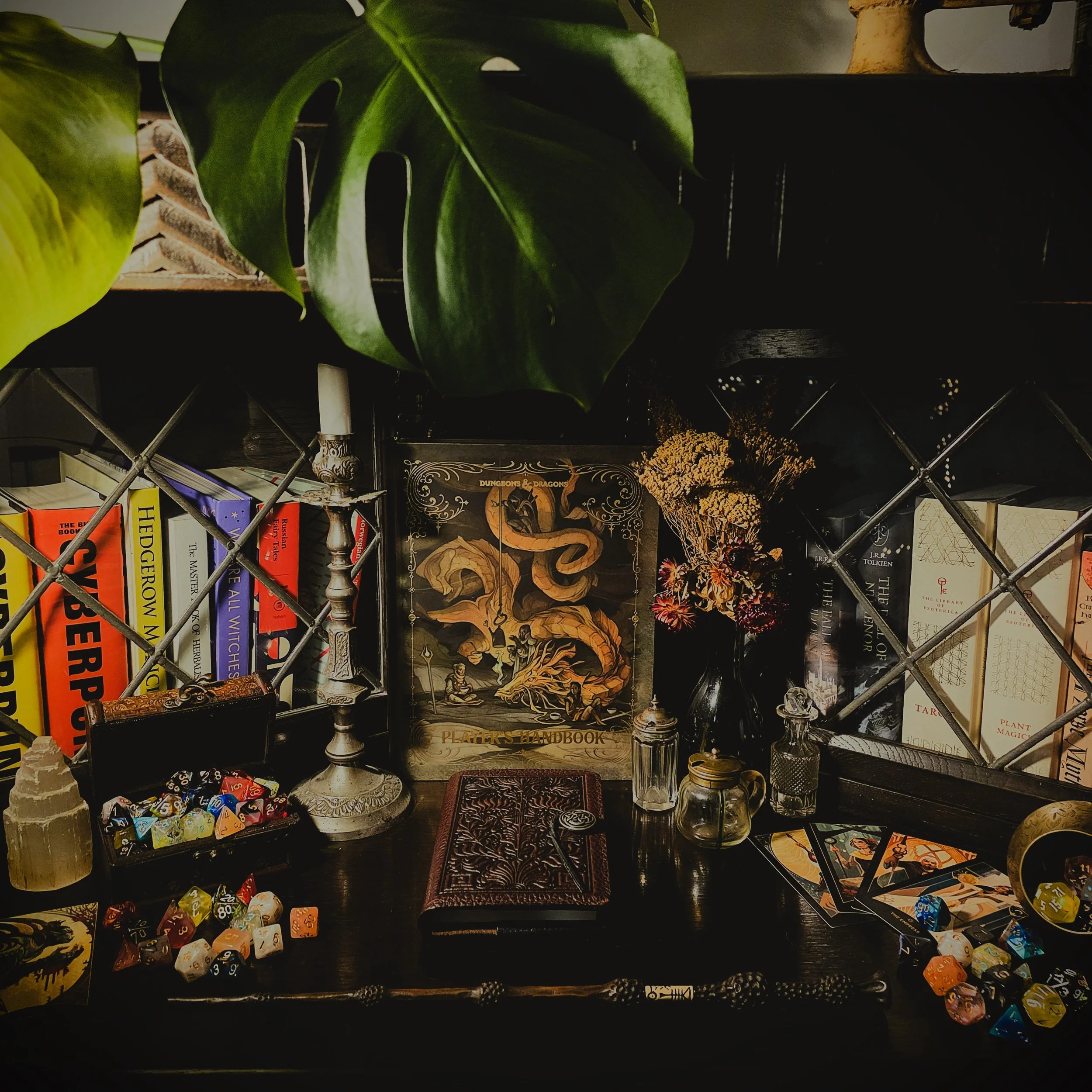 A tabletop setup with books, game components, a candle holder, and decorative items. A large Monstera leaf partially covers the top of the image. There are dice scattered on the table and books about fantasy and role-playing games. A dark vase with dried flowers, a closed leather-bound journal or notebook, and various small jars or bottles are also visible.