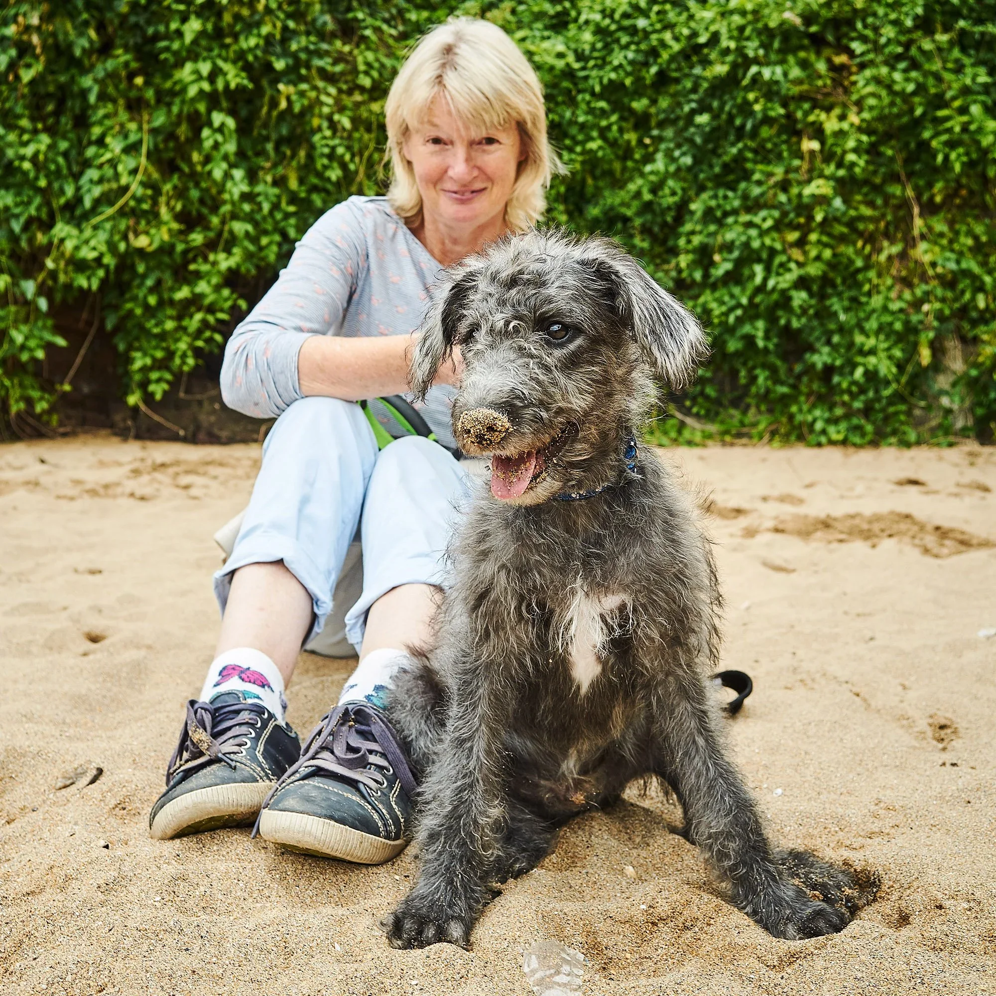 A woman sitting on sand with a muddy beach and green bushes in the background, smiling at a wet, muddy, grey and black puppy sitting in front of her.