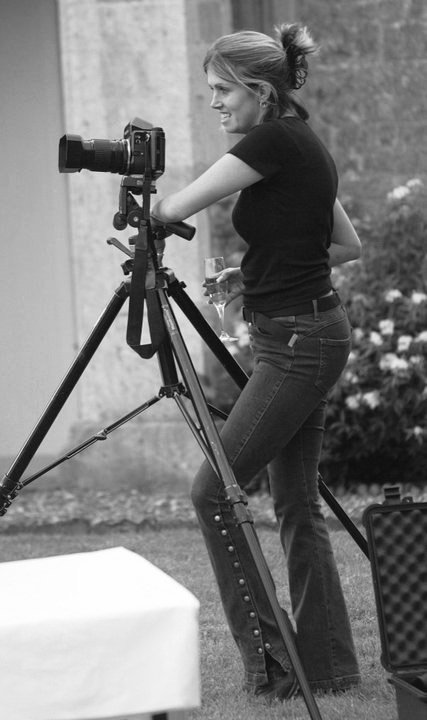 A woman operating a camera on a tripod outdoors, holding a glass of wine, smiling, with a garden and stone wall in the background.