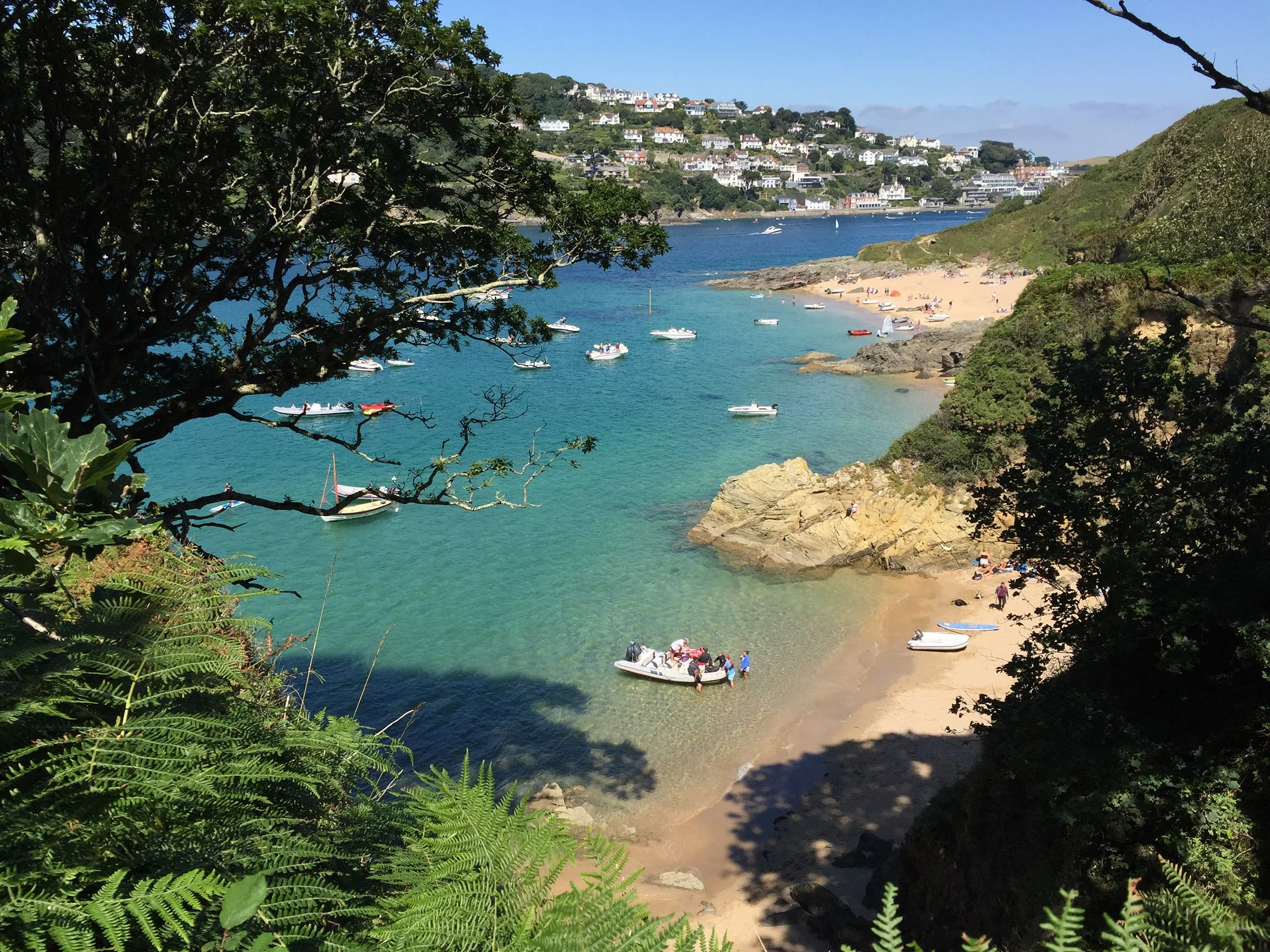 A scenic view of a beach with boats in the water, surrounded by green trees and hills, with a town in the background.
