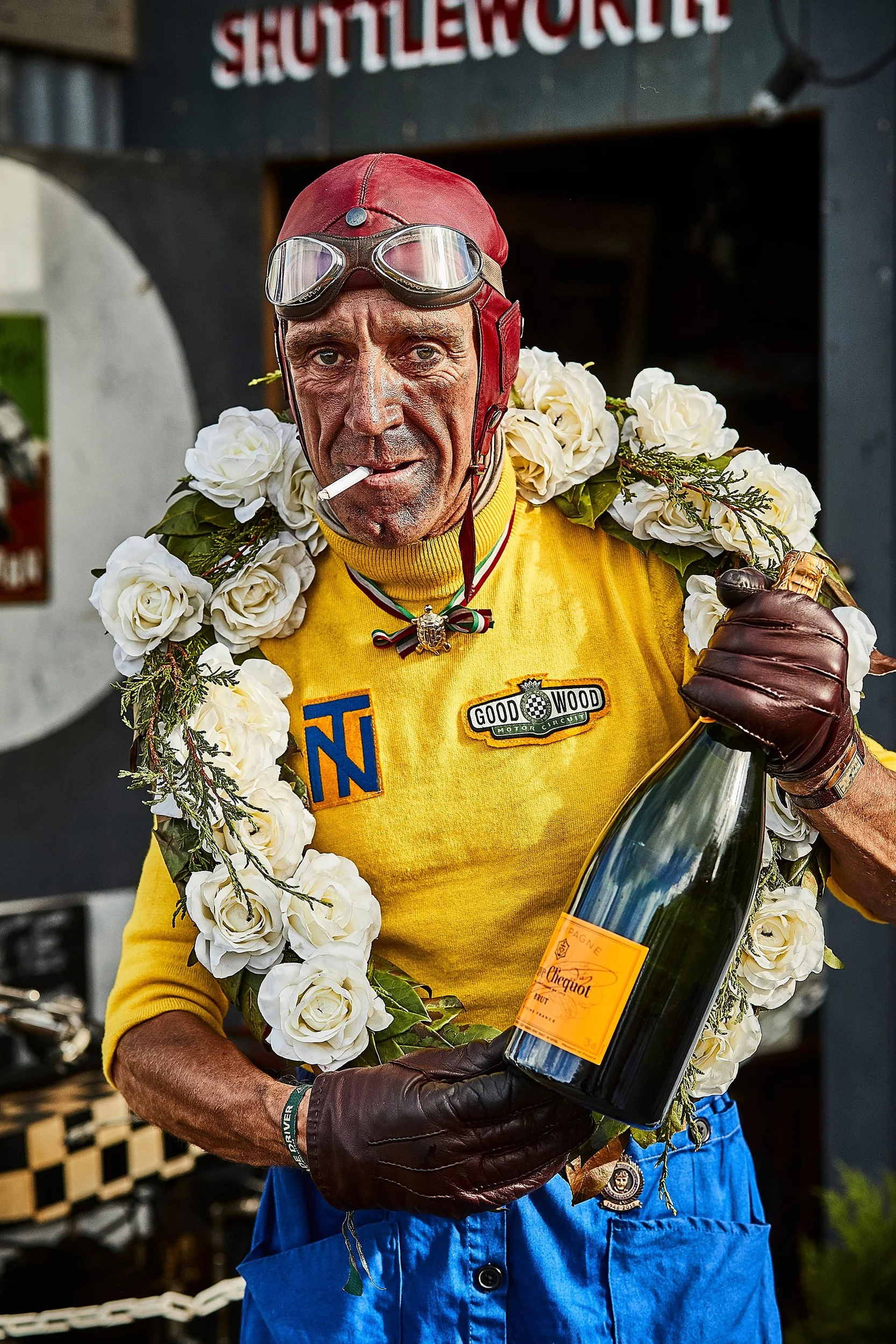 An elderly man wearing a red leather helmet with goggles, a yellow jacket, and a garland of white roses around his neck. He is holding a bottle of champagne and has a cigarette in his mouth. The man appears to be at a celebratory event or gathering.