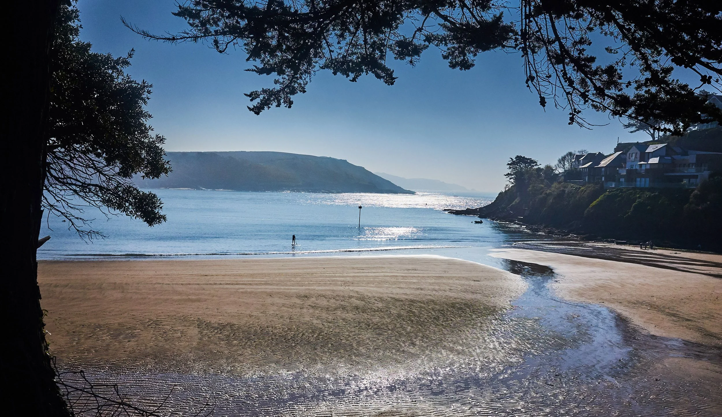 A peaceful beach scene with a sandy shoreline, a calm ocean, and a distant hillside. Tree branches frame the top of the image, with houses on a hill to the right. The sun reflects on the water, and a person is walking along the shore.