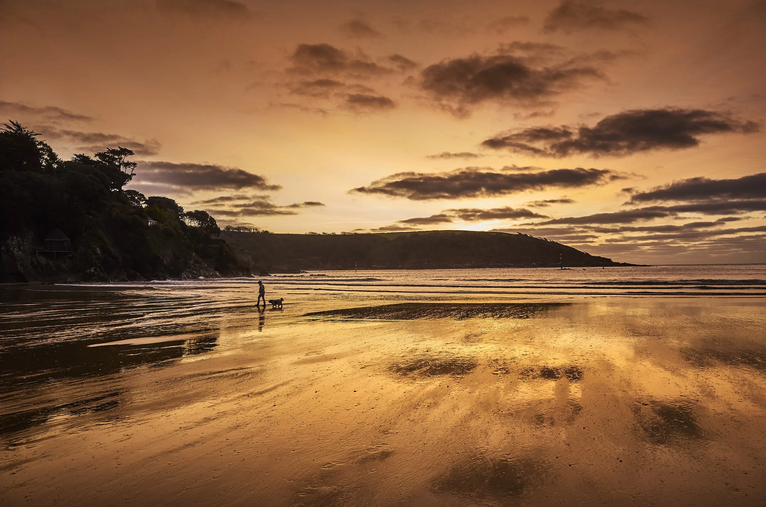 Sunset over a beach with a person and dog walking along the shore, reflection of the sky on wet sand, rocky coastline on the left, and distant land on the horizon.