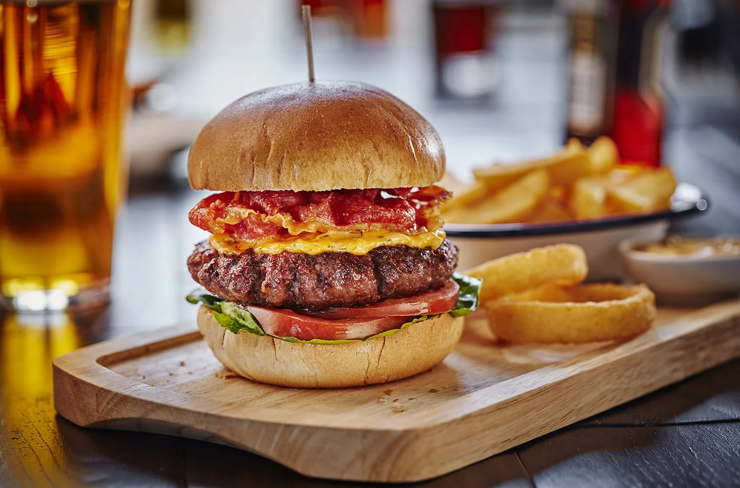 A close-up of a cheeseburger on a wooden tray with onion rings and French fries in the background. The burger has a bun, tomato, lettuce, a beef patty, melted cheese, crispy bacon, and scrambled eggs.