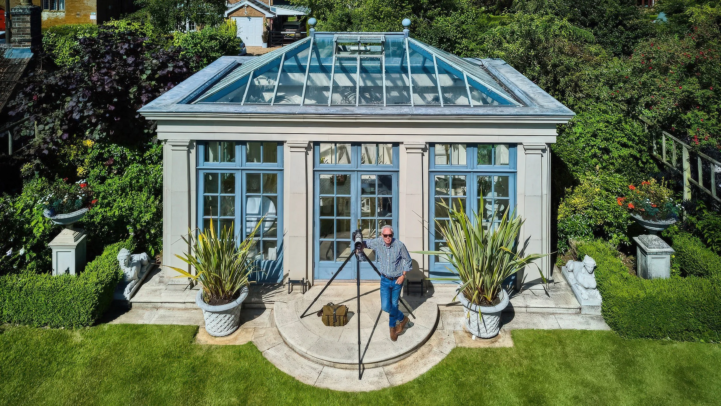 A man wearing sunglasses and a checkered shirt stands in front of a glass greenhouse, holding a telescope on a tripod outside the greenhouse. The greenhouse has large blue windows and is decorated with potted plants and statues, with a lush garden and residential houses in the background.