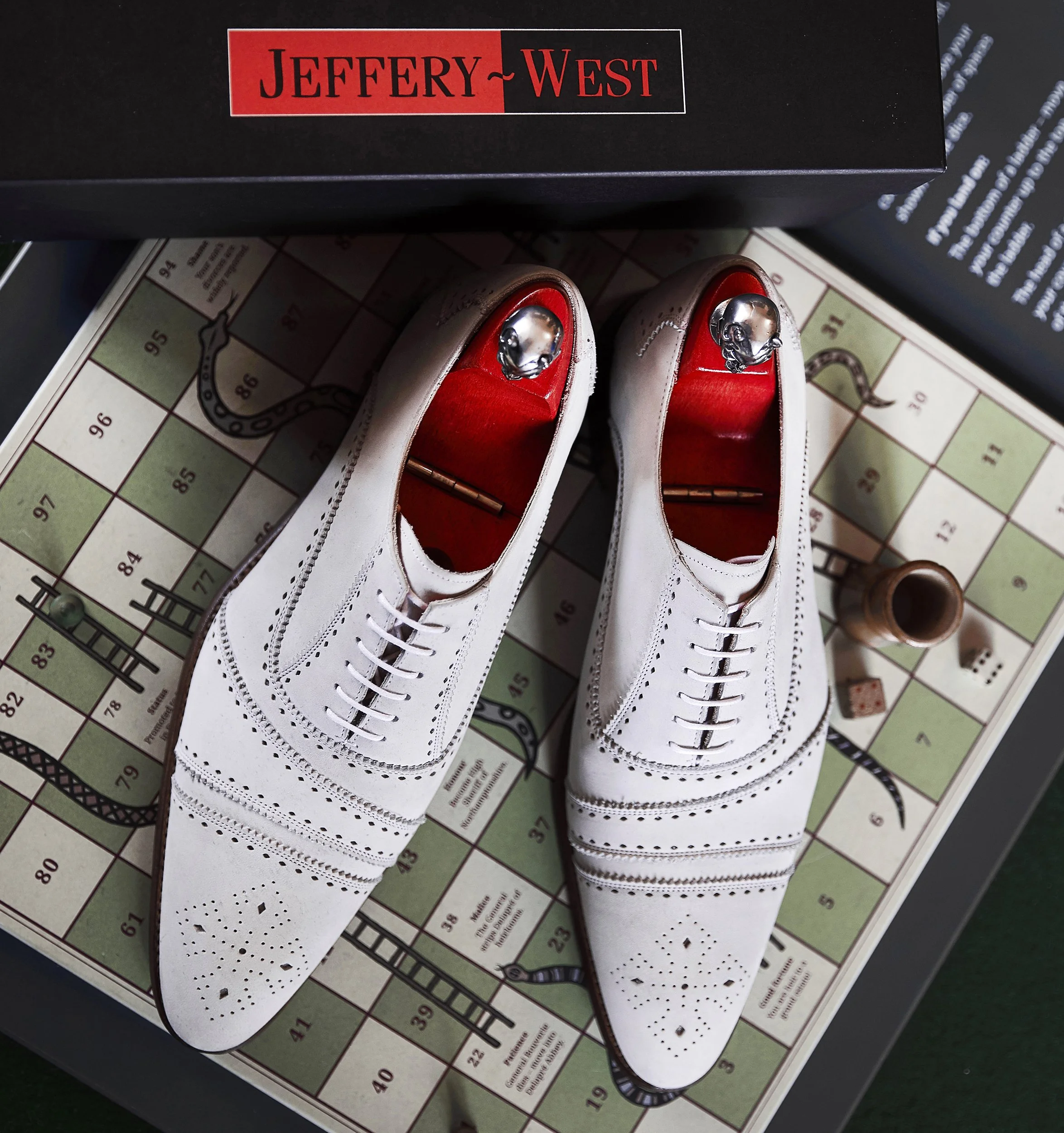 Pair of white leather dress shoes with brogue details and silver toe caps, placed on a vintage Monopoly game board. A black box with red and black text reading 'Jeffery~West' is partly visible at the top.