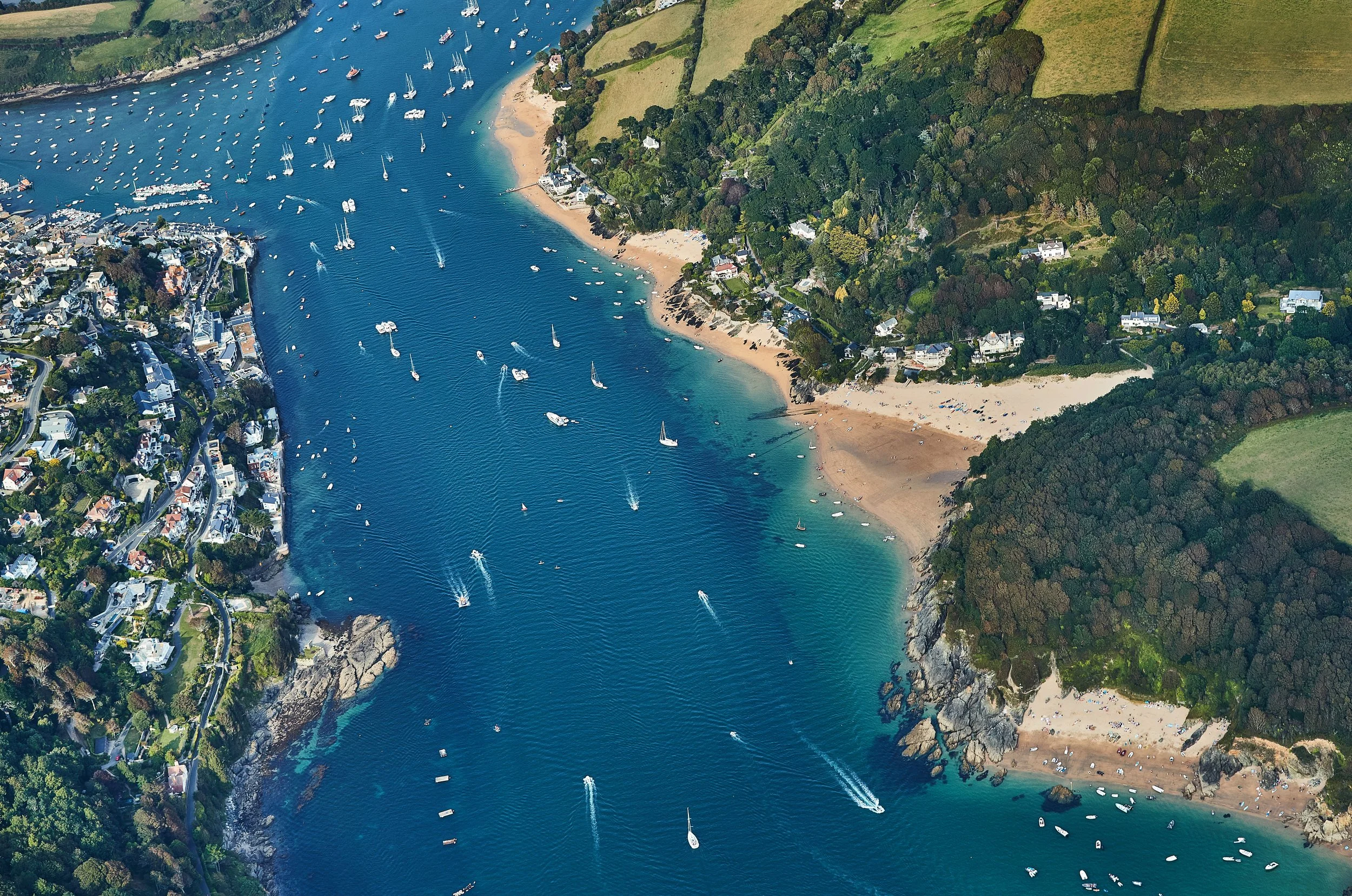 Aerial view of a coastal area with a deep blue bay filled with boats and sailboats, sandy beaches, and green hills with houses.