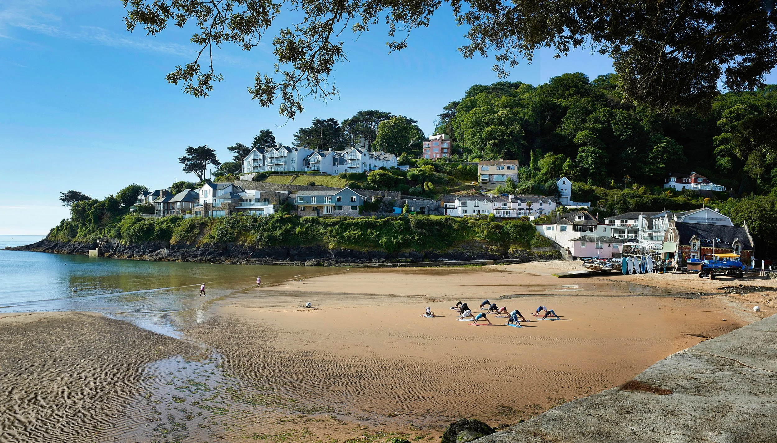 People practicing yoga on the sandy beach with houses on a hill in the background and trees overhead.
