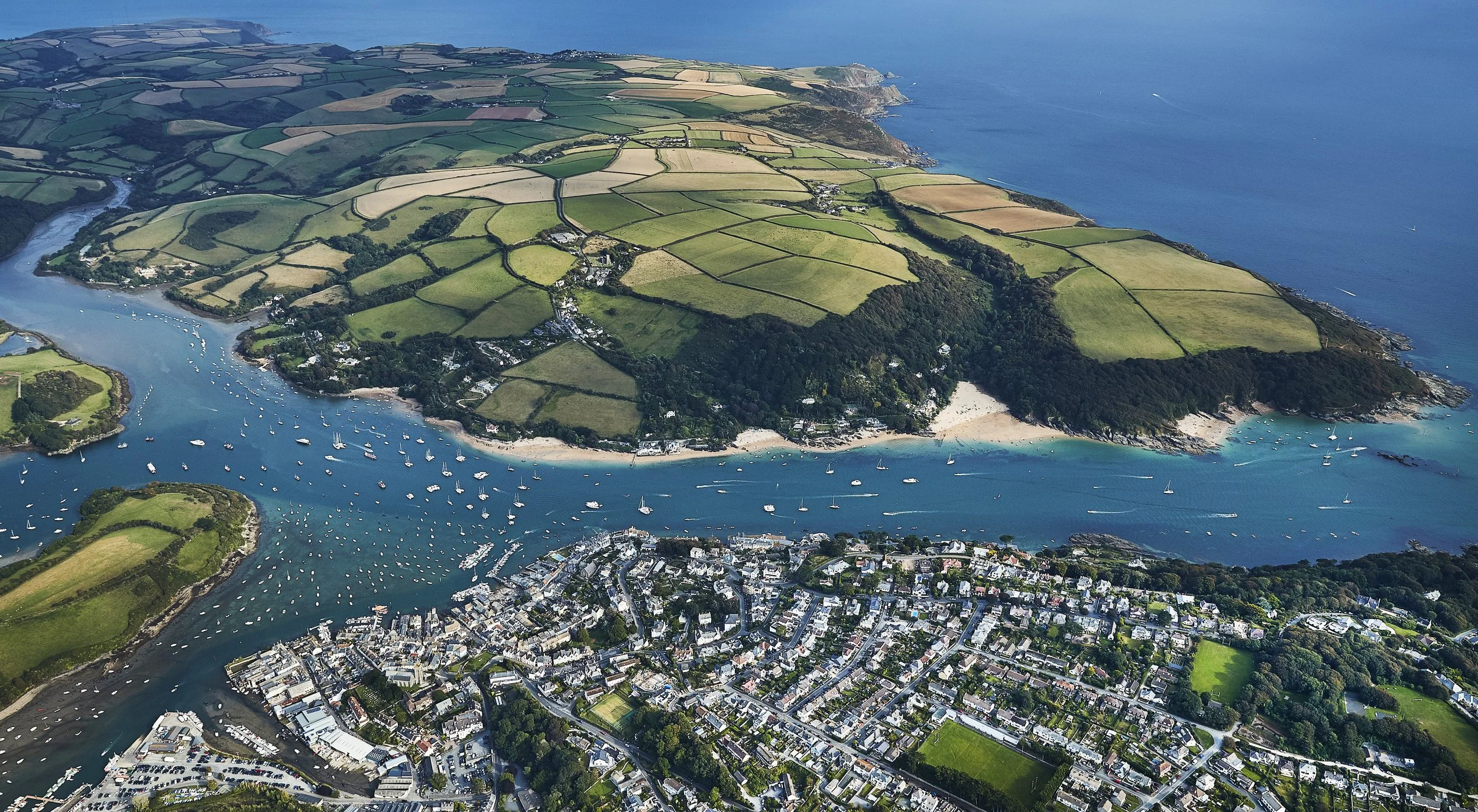 Aerial view of a coastal town with a river, boats, green fields, and a surrounding landscape of hills and ocean.