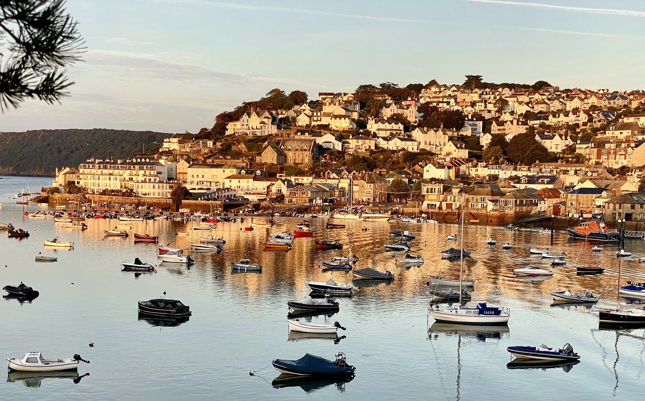 A coastal village with numerous boats moored in calm water during sunset, overlooking a hillside with closely packed houses and buildings.