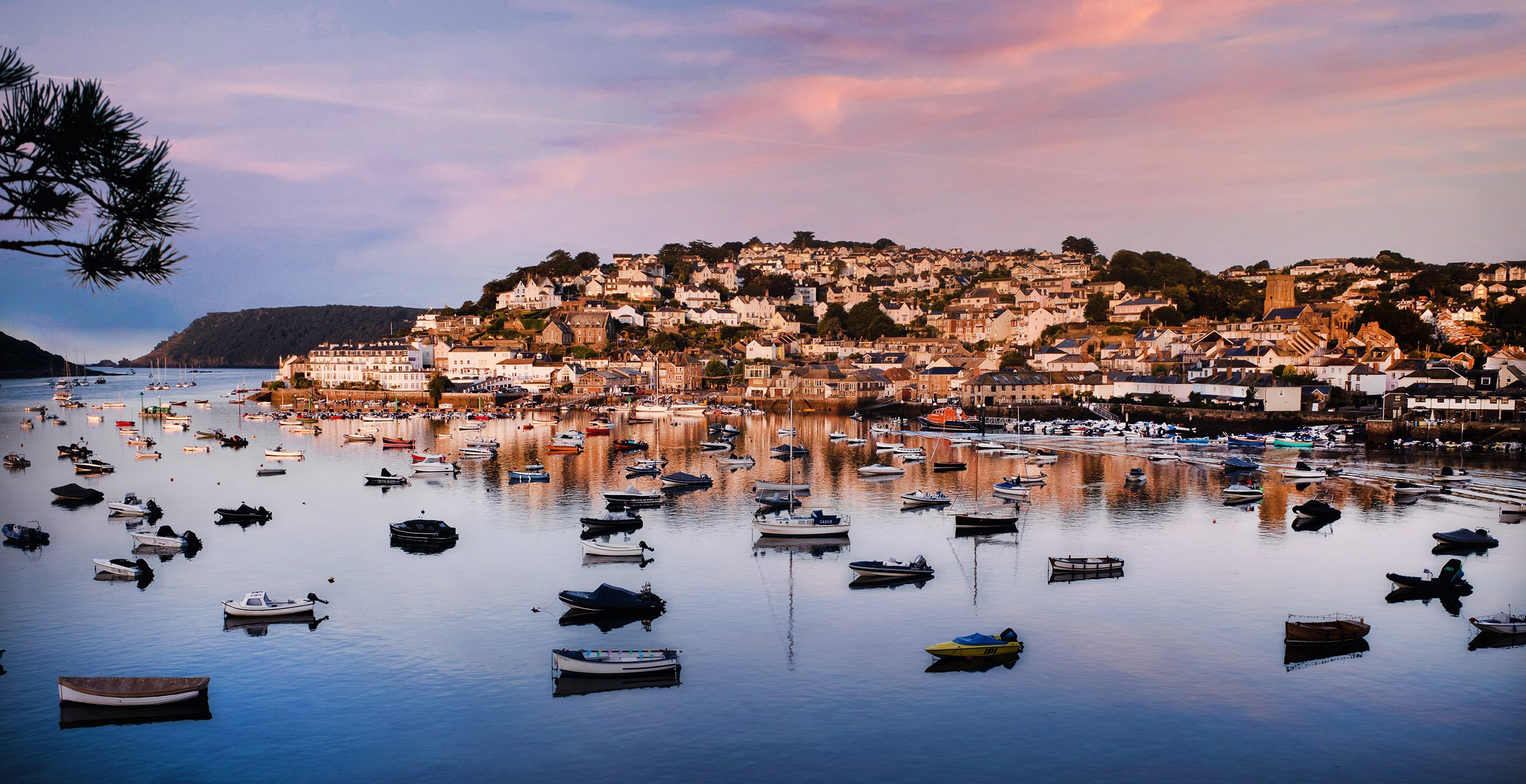 A harbor at sunset with numerous boats floating on calm water and houses on a hillside in the background
