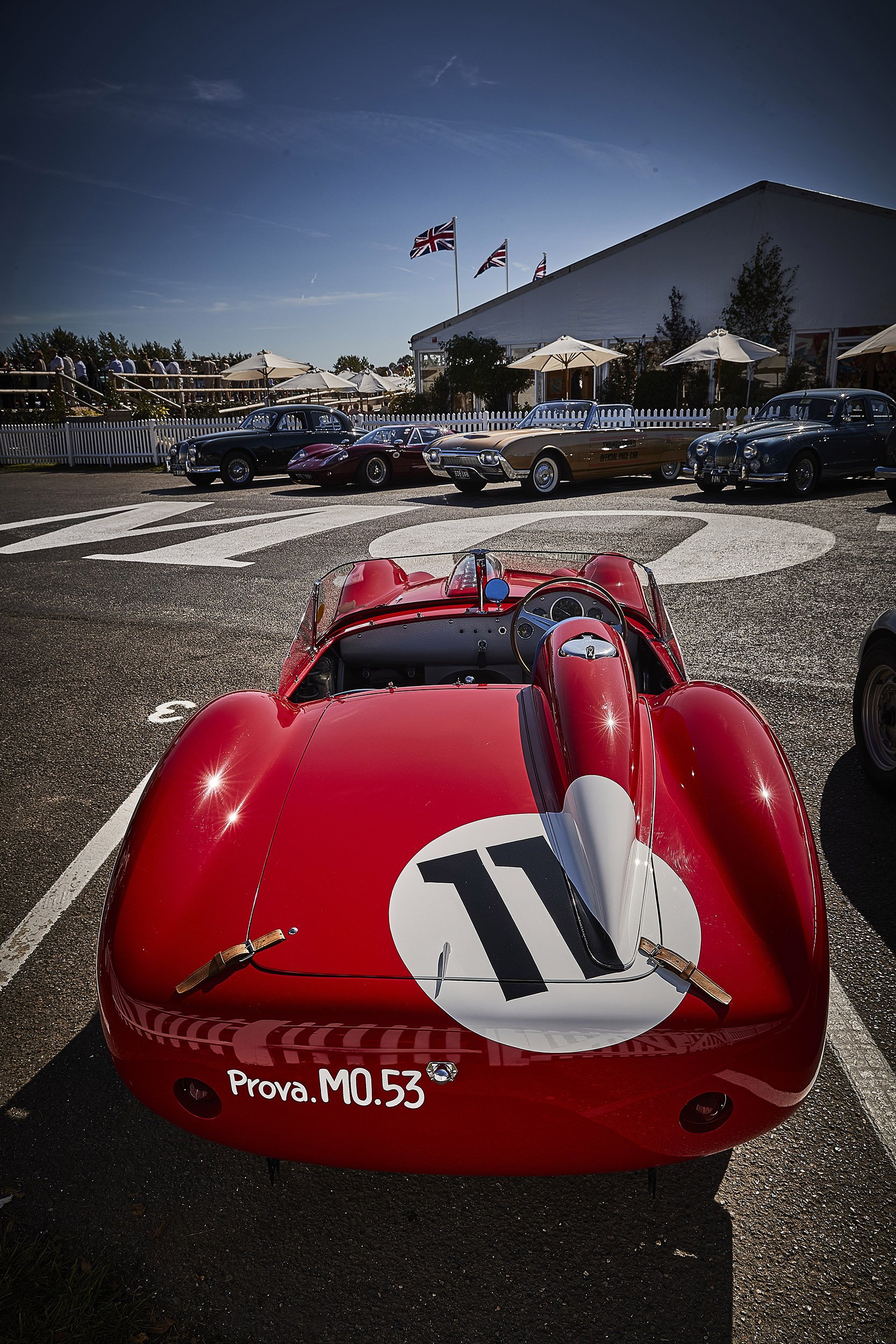 A row of vintage race cars parked at a concours event, with a red open-wheel racing car in the foreground and classic cars in the background under a clear blue sky.