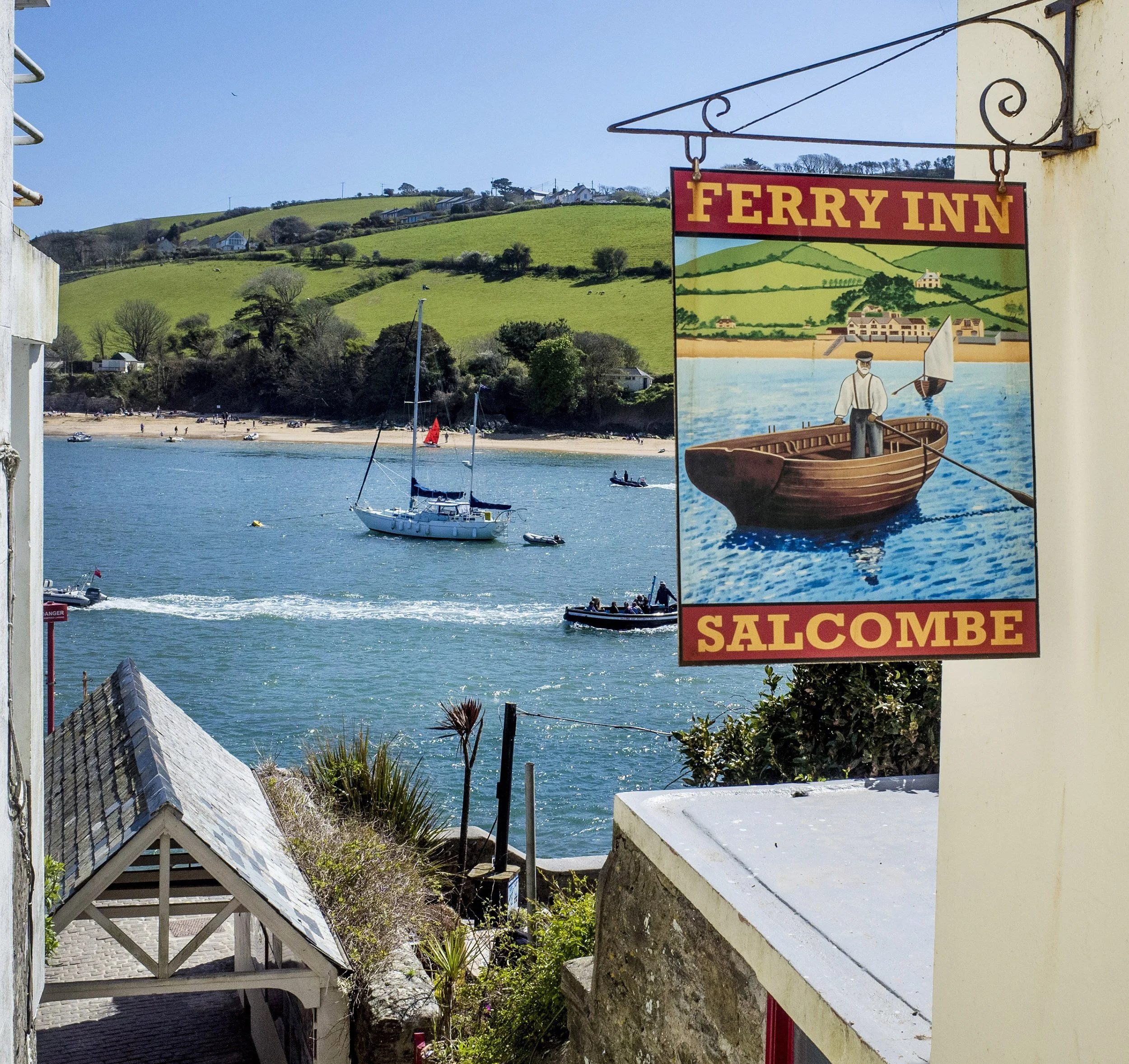 A coastal scene with boats on water, a beach, green hills in background, and a sign reading 'Ferry Inn Salcombe' depicting a boat with a person steering.