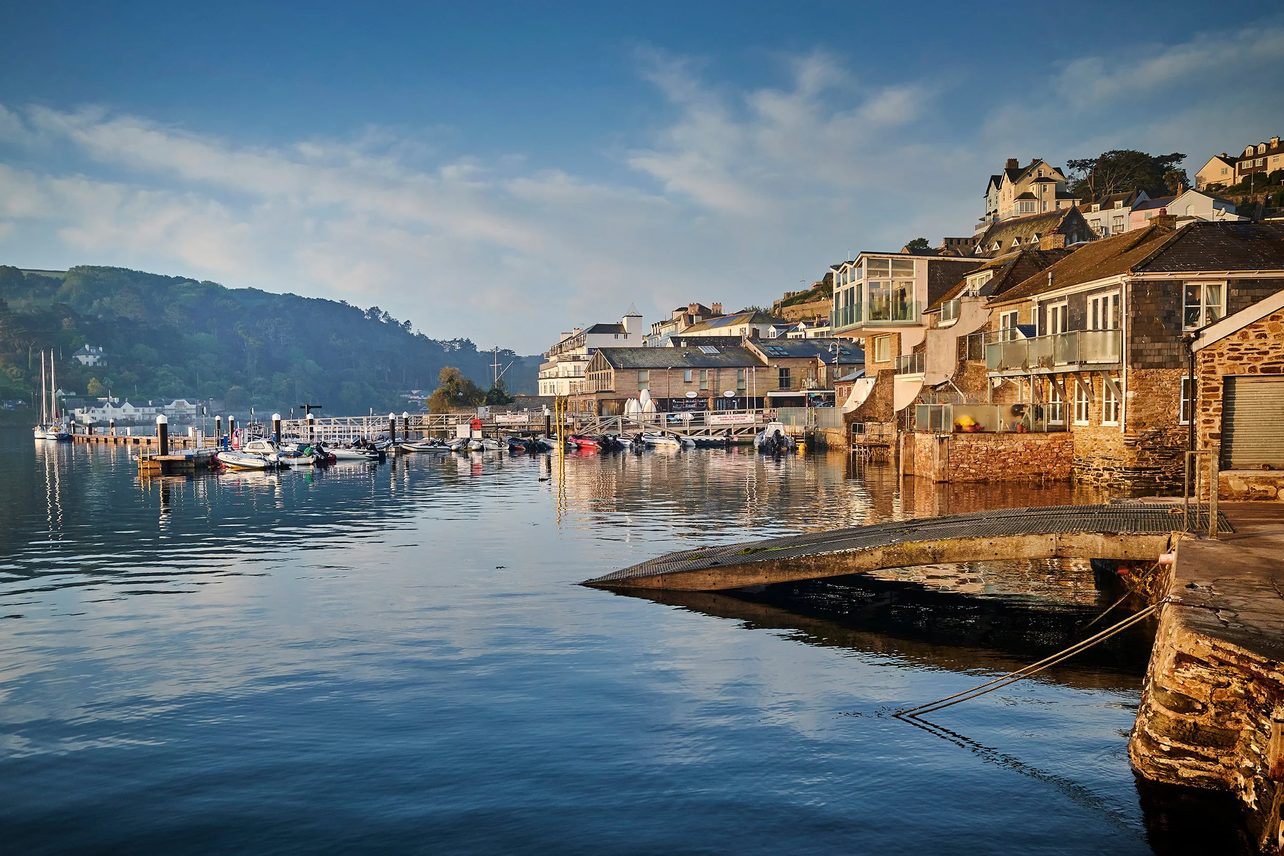 A scenic harbor with boats docked along a pier, surrounded by colorful houses on a hillside, under a partly cloudy sky.