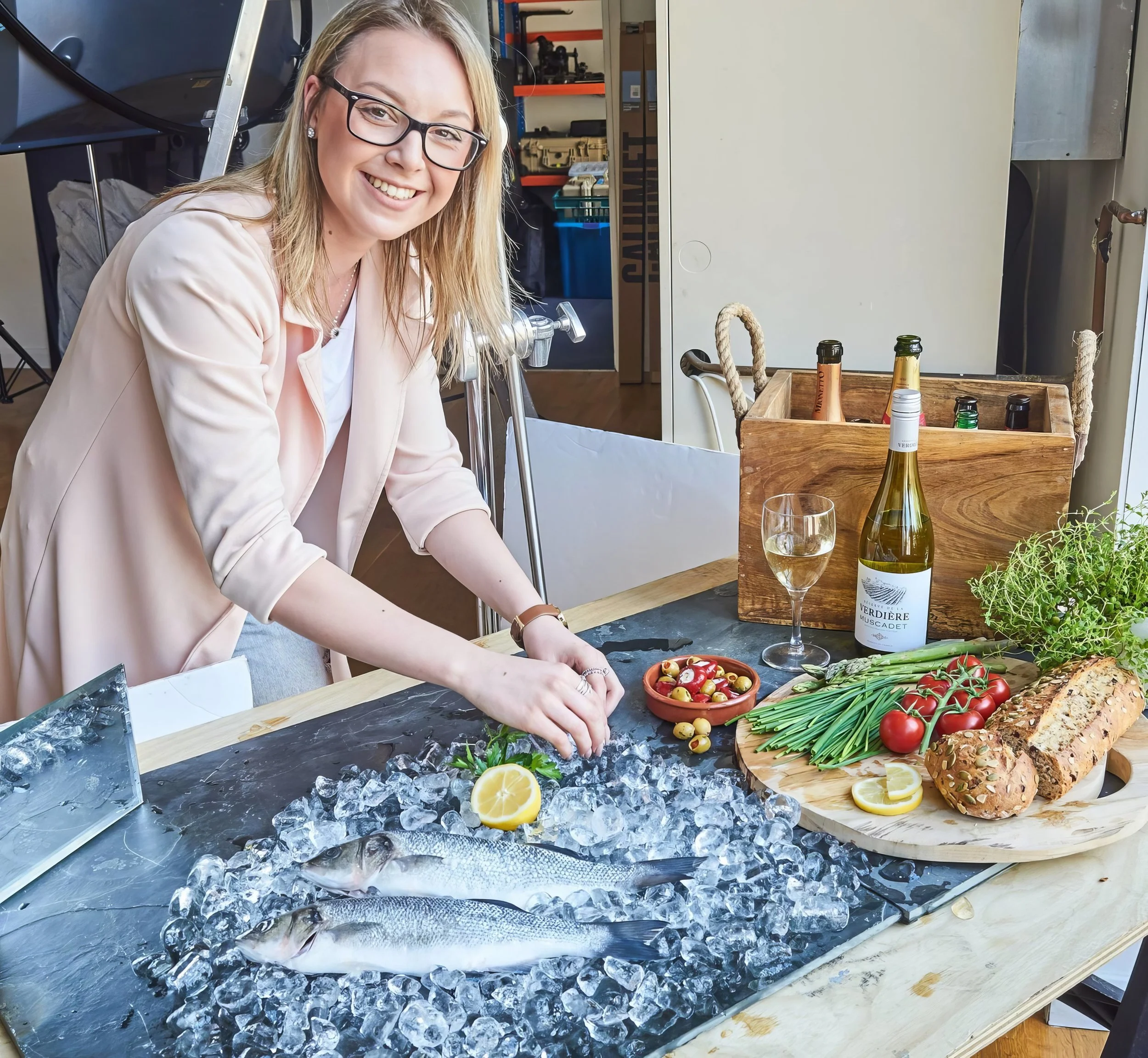 A woman with glasses and blonde hair smiling while preparing fresh fish on a bed of ice, with lemons, tomatoes, herbs, bread, chips, wine, and a bowl of small tomatoes on the table.