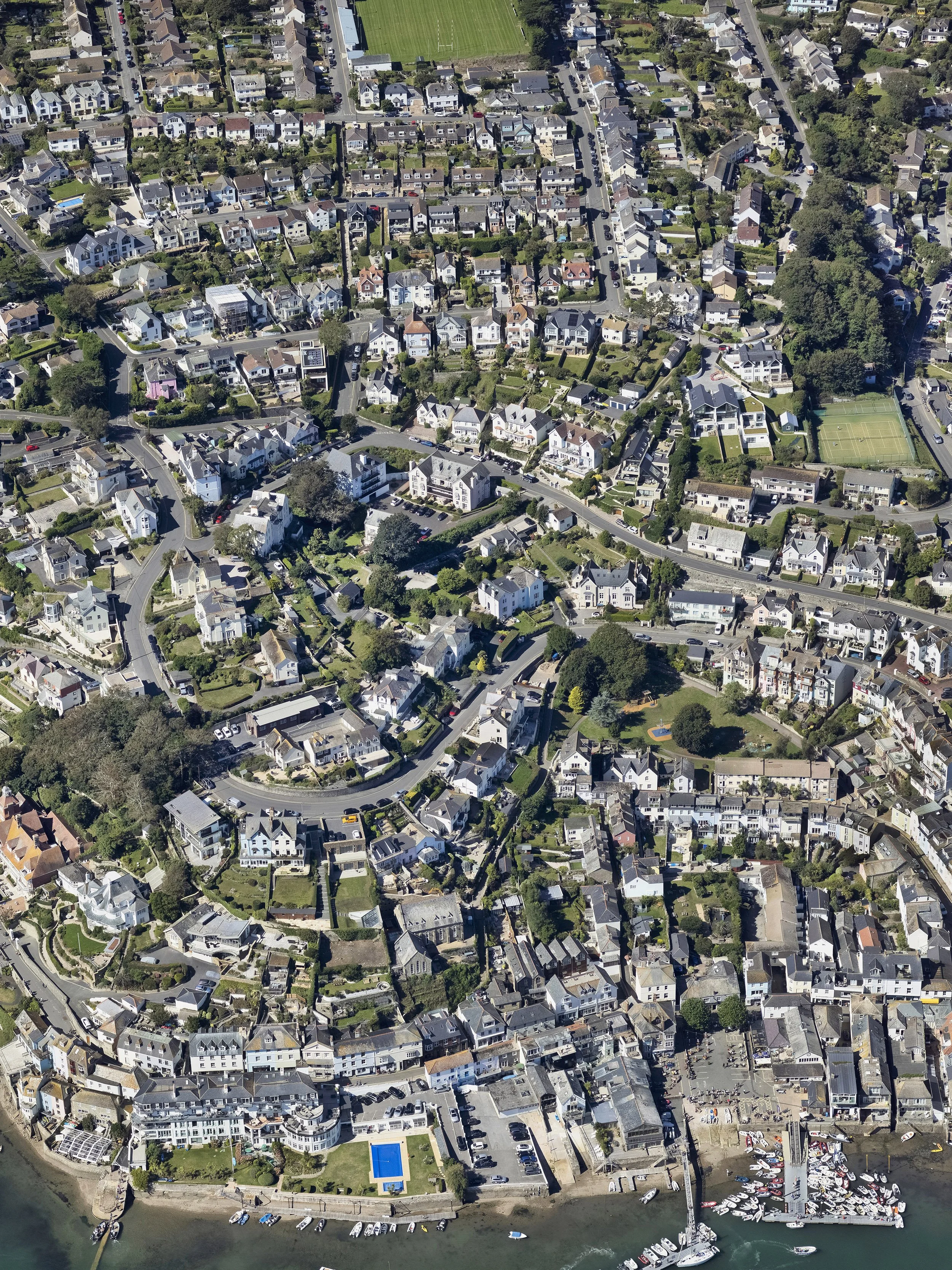 Aerial view of a coastal residential neighborhood with houses, green spaces, tennis courts, a marina with boats, and a body of water at the bottom.