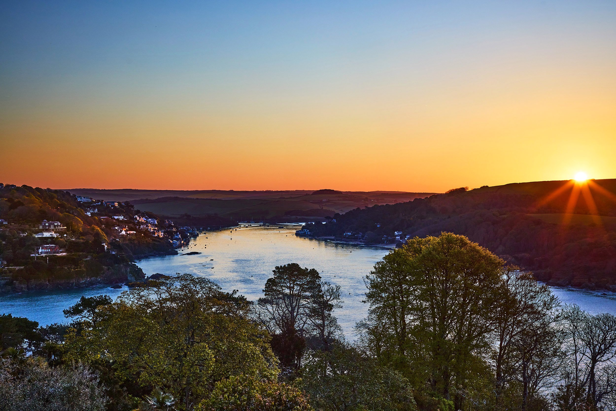 Sunset over a river with trees in the foreground and houses along the hillsides on both sides of the river.