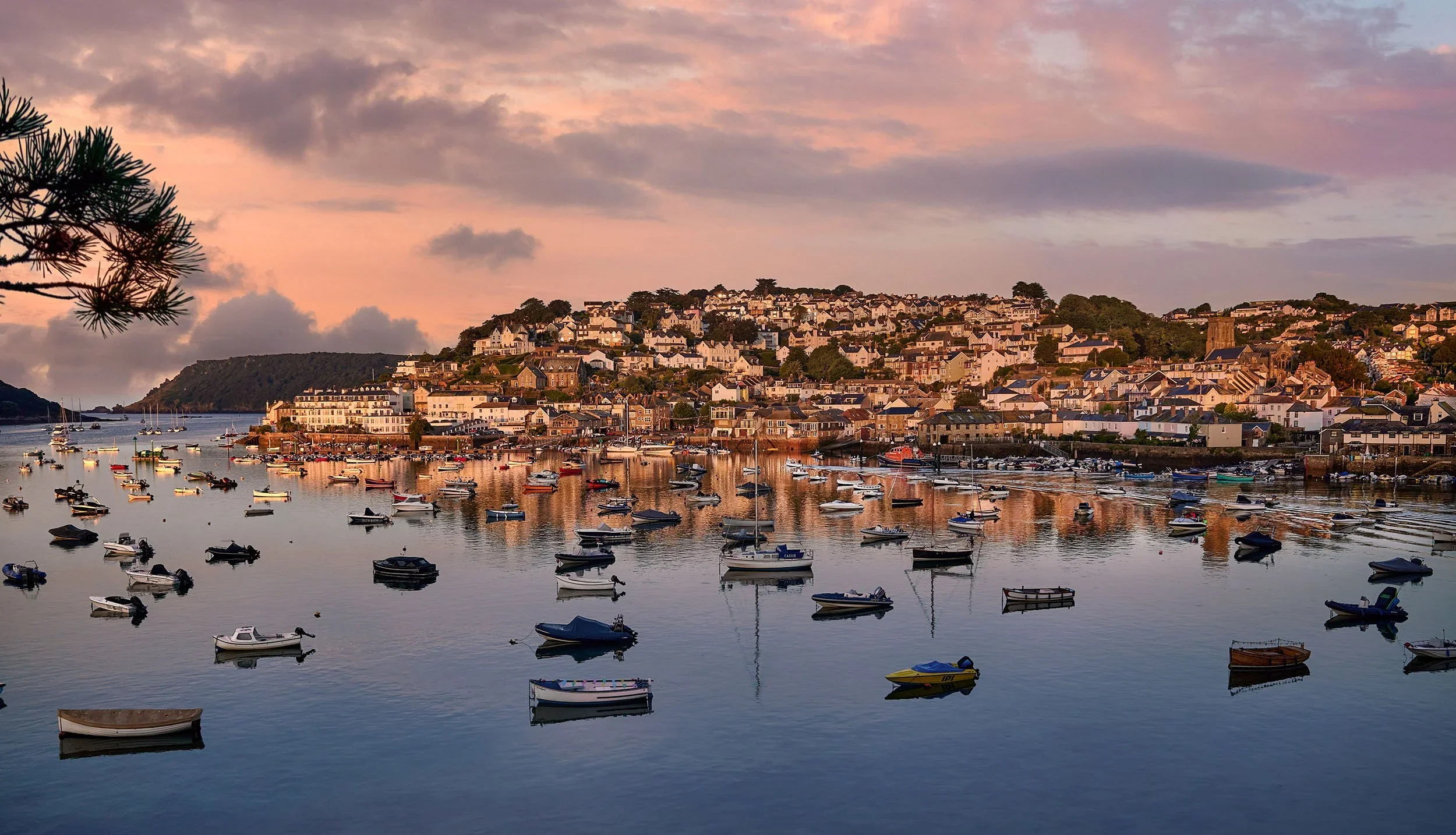 A harbor at sunset with boats floating on calm water, a hillside town with many houses and a church, and a sky with pink and purple clouds.