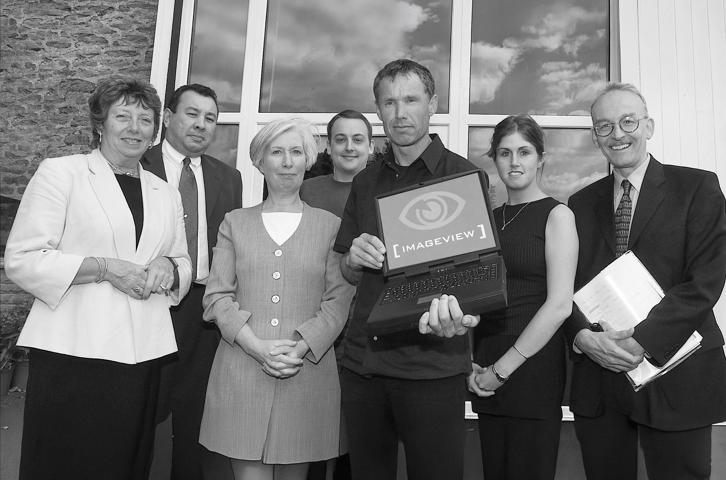 A group of eight professionally dressed individuals standing together inside in front of a large window. One person in the center holds an open laptop displaying the 'ImageView' logo.