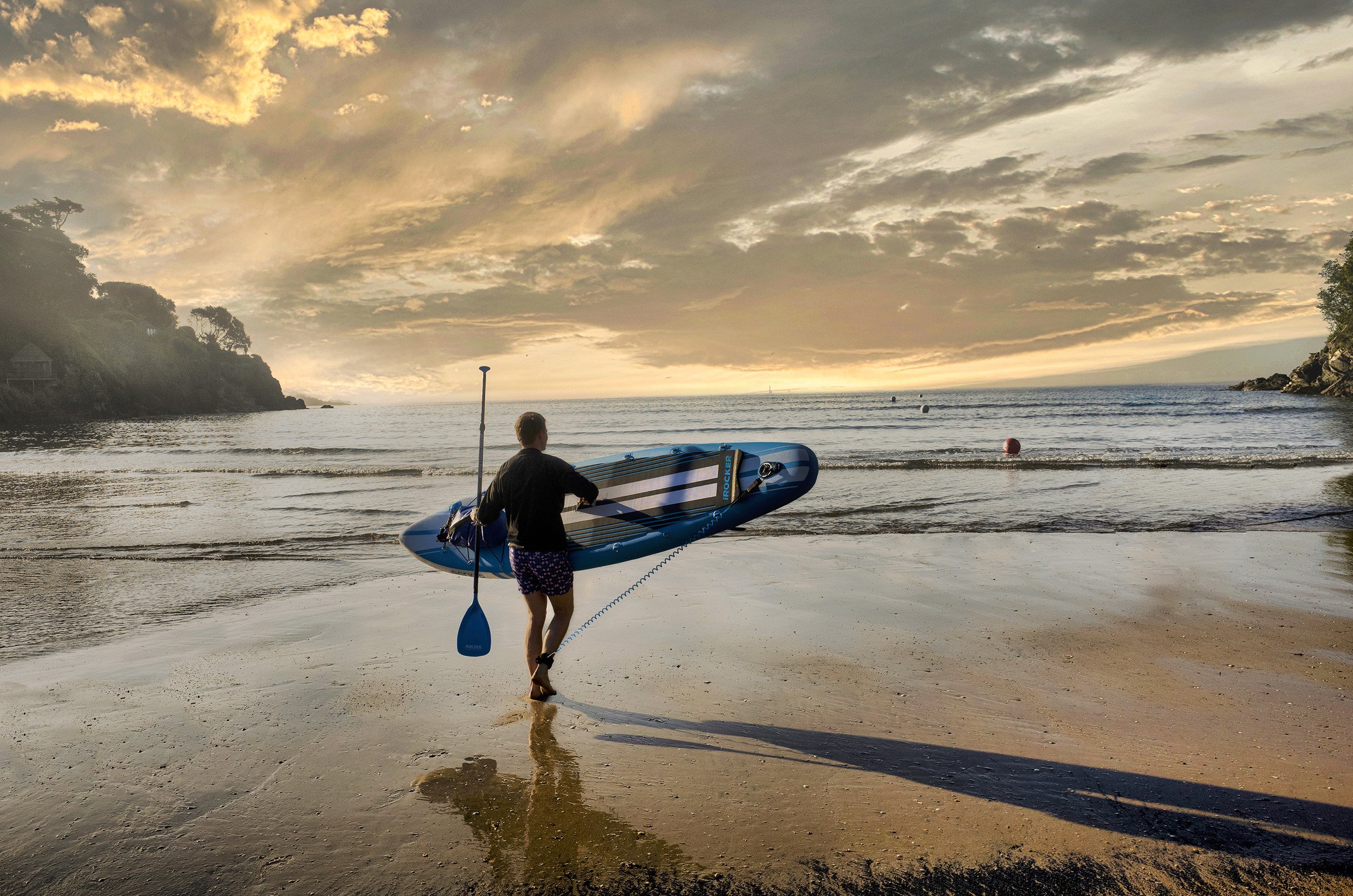 Person carrying a paddleboard and paddle walking on a sandy beach at sunset, with calm ocean water and a cloudy sky.