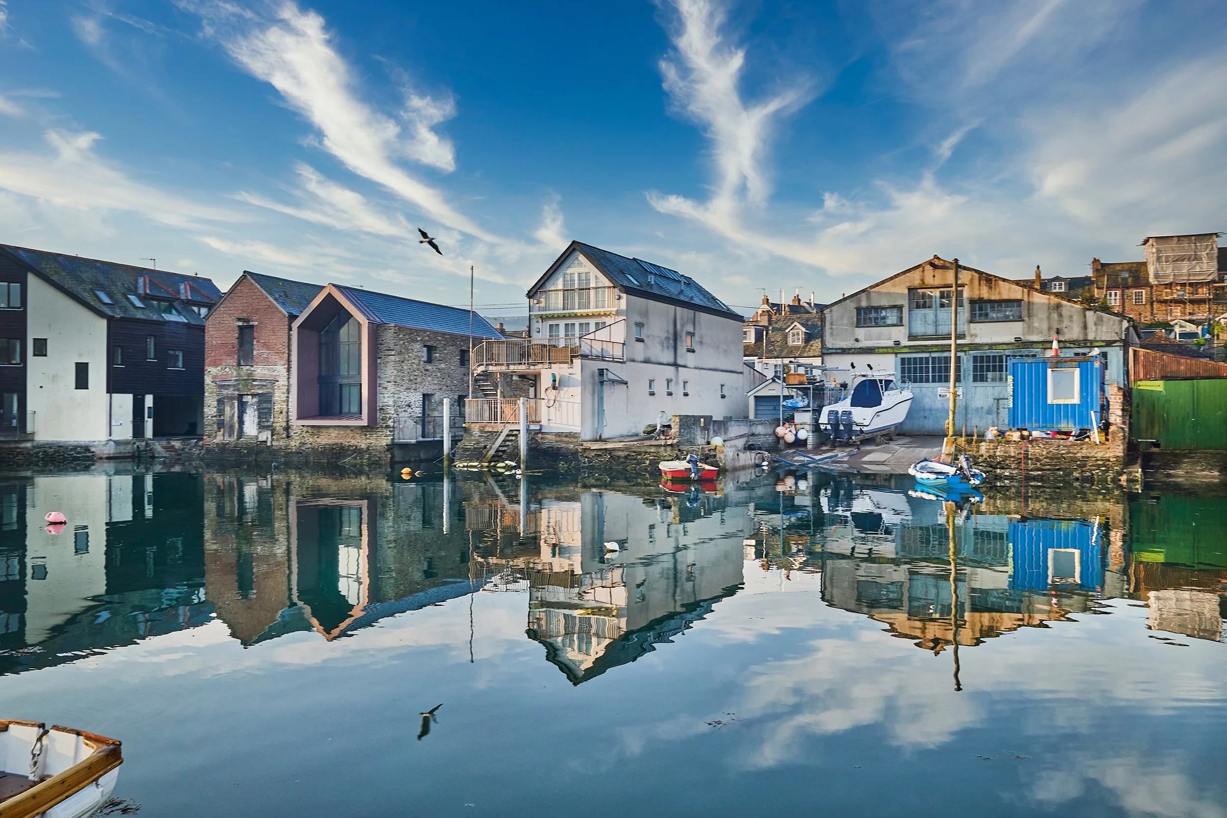 A harbor scene with calm water reflecting colorful boats, houses, and the blue sky with wispy clouds.