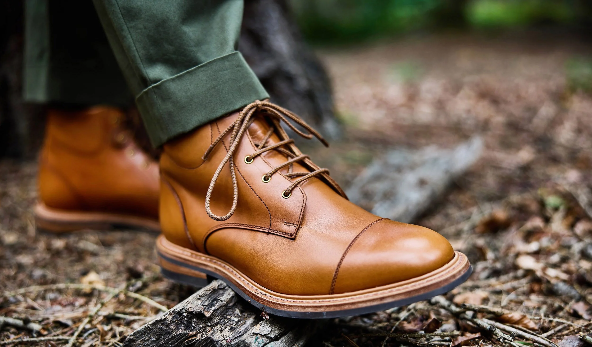 Close-up of a person's brown leather hiking boot on a fallen tree branch in a forest, with green pants and blurred background.