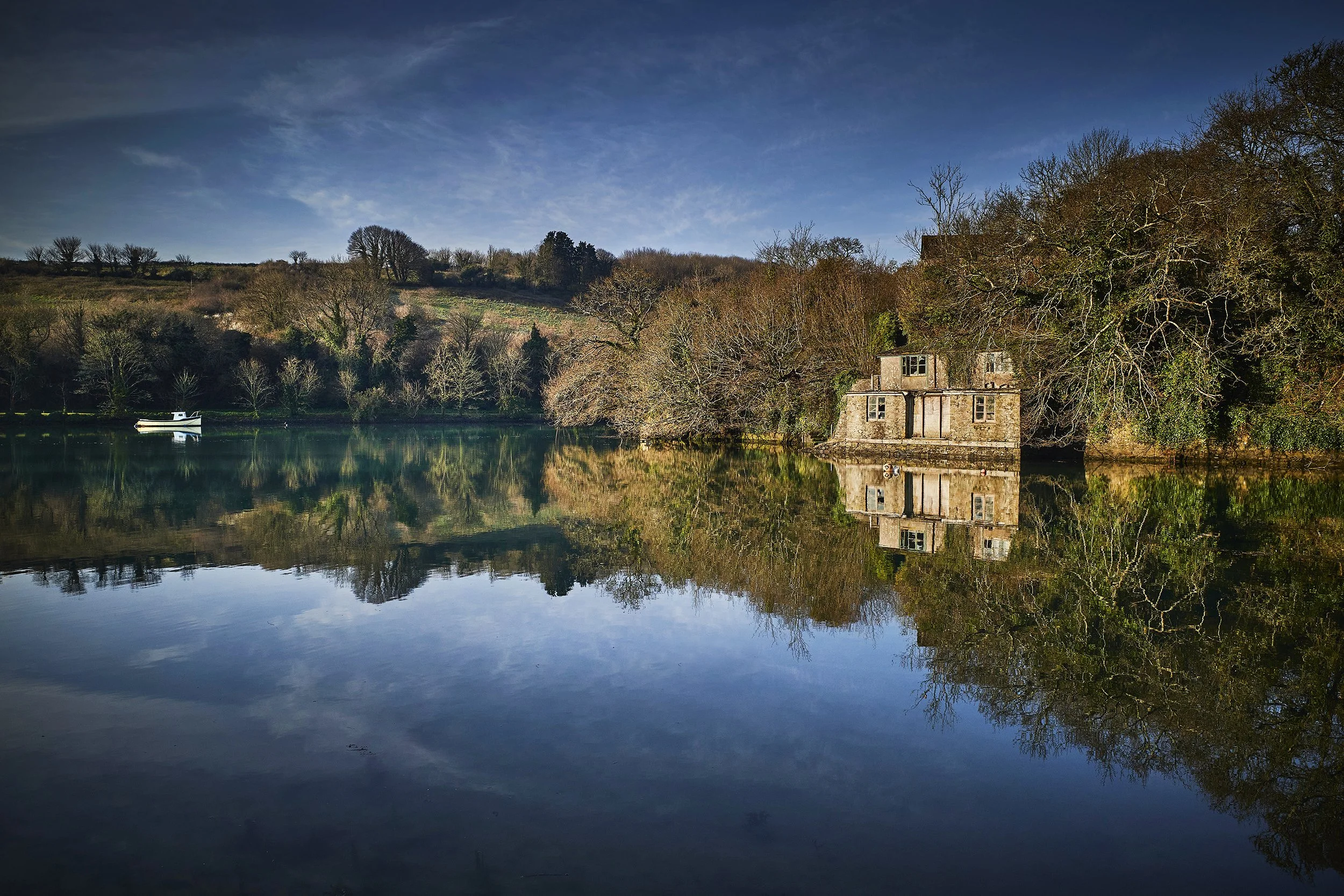 A serene landscape with a body of water reflecting a small house and trees on the shoreline, under a blue sky with clouds.