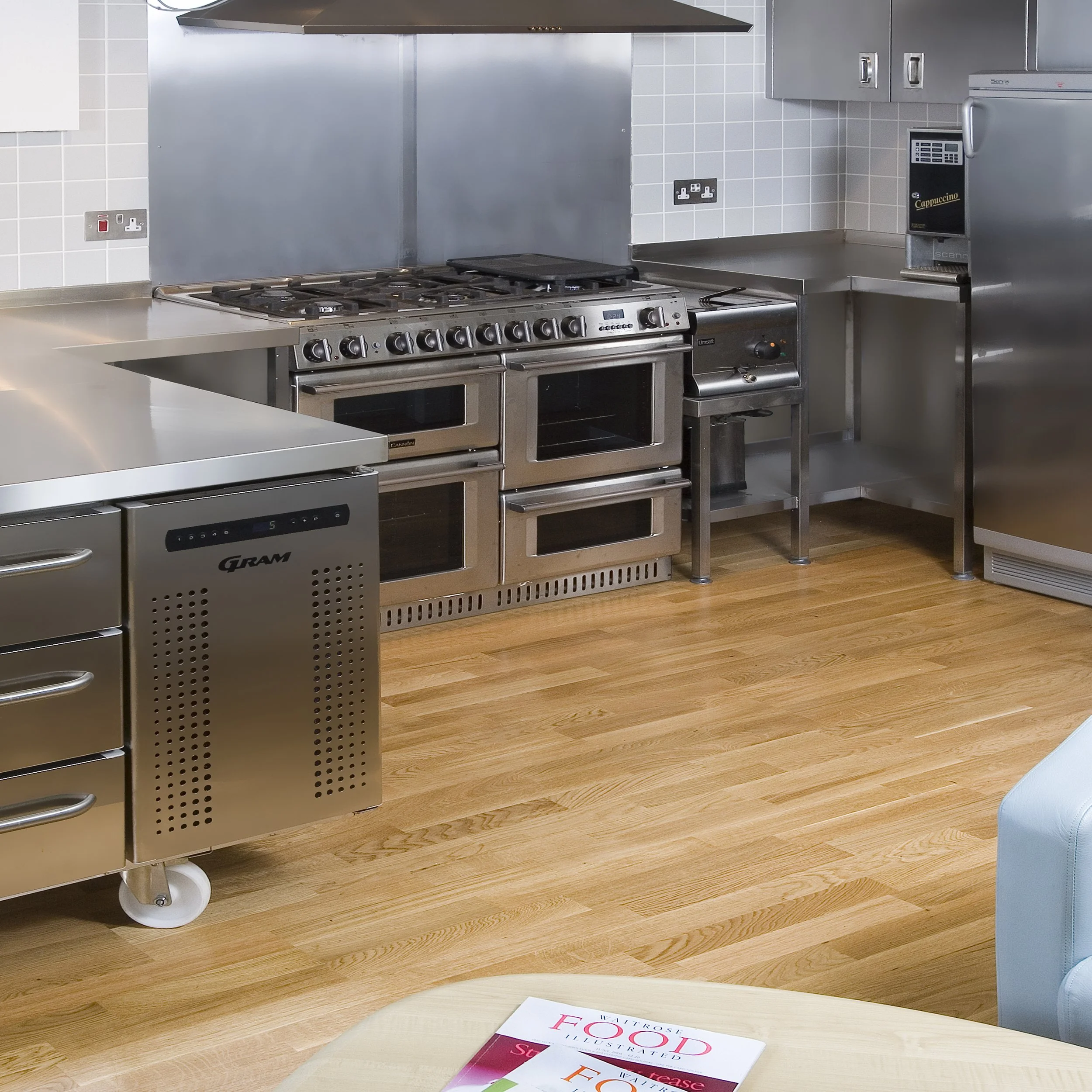 Commercial kitchen with stainless steel appliances, including a stove, oven, refrigerator, and a coffee machine, with a wooden floor and a book on a table in the foreground.