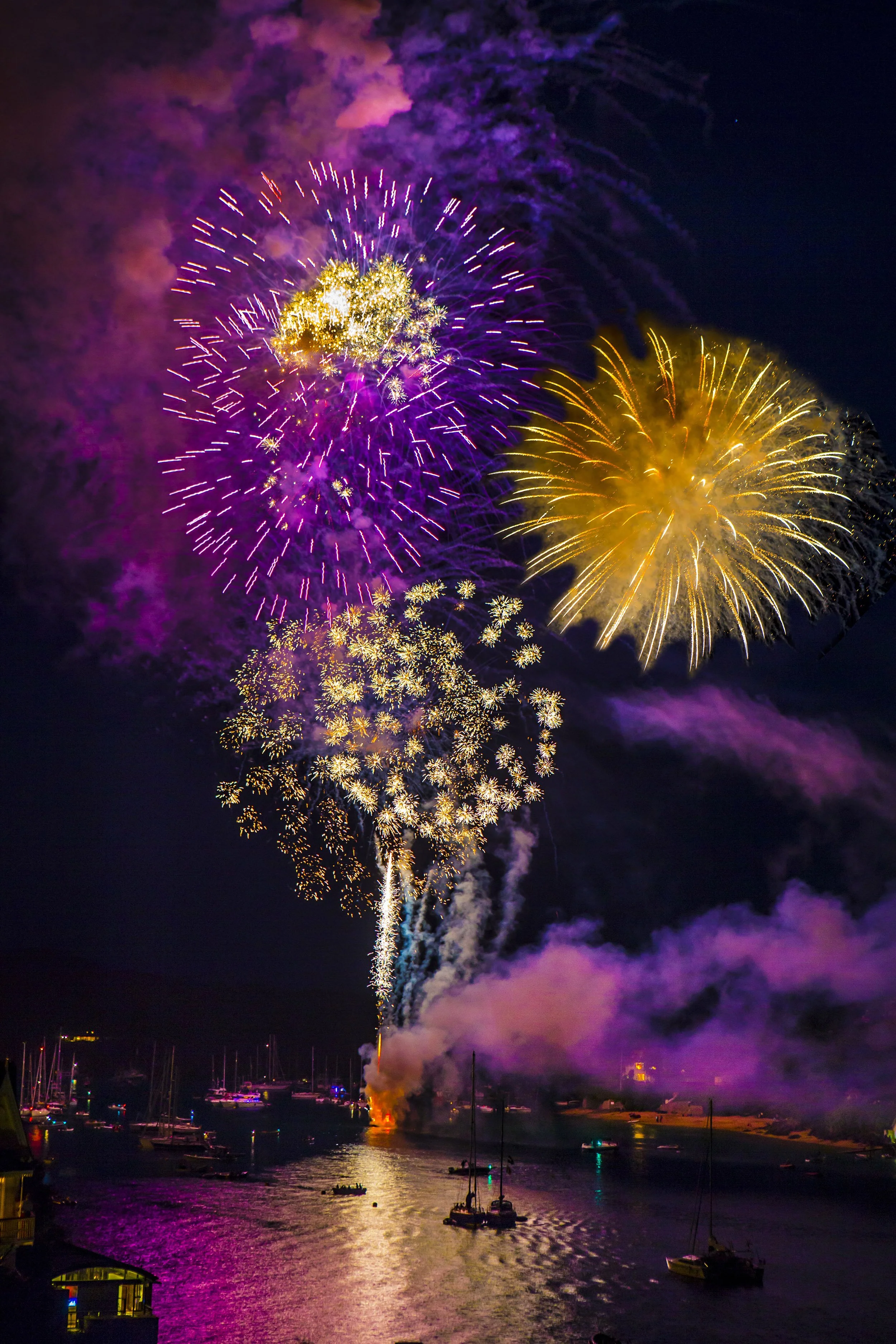 Colorful fireworks display over a body of water at night, with boats and reflections visible below.