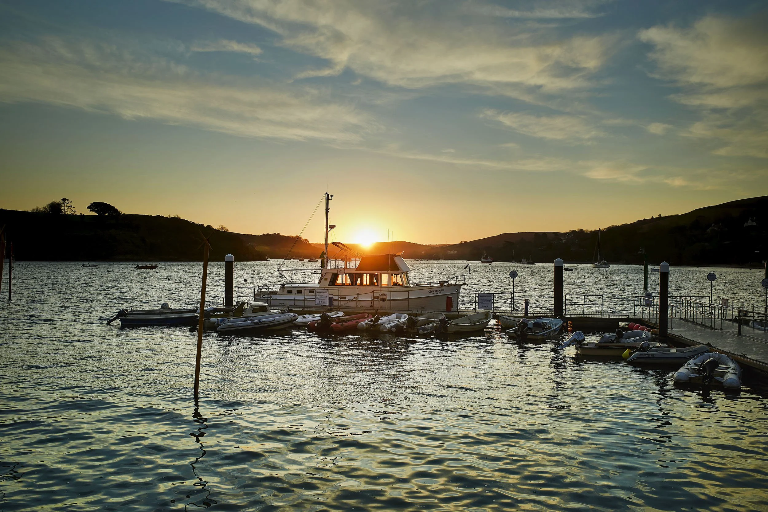 Boats docked at a marina during sunset with a hillside in the background.