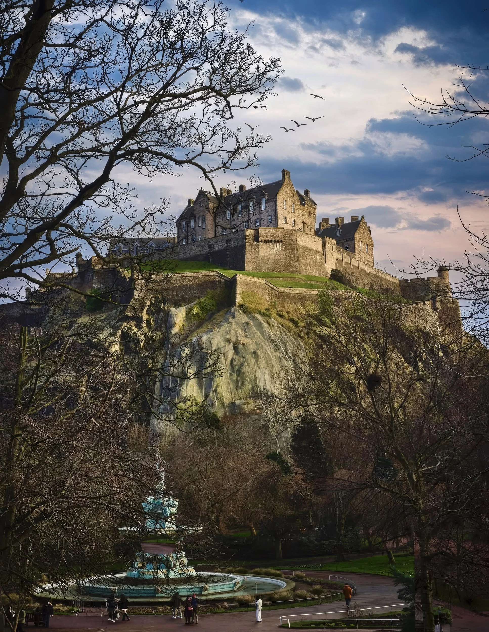 A historic castle atop a hill, with leafless trees and a cloudy sky in the background. In the foreground, a park with a fountain and visitors walking around.