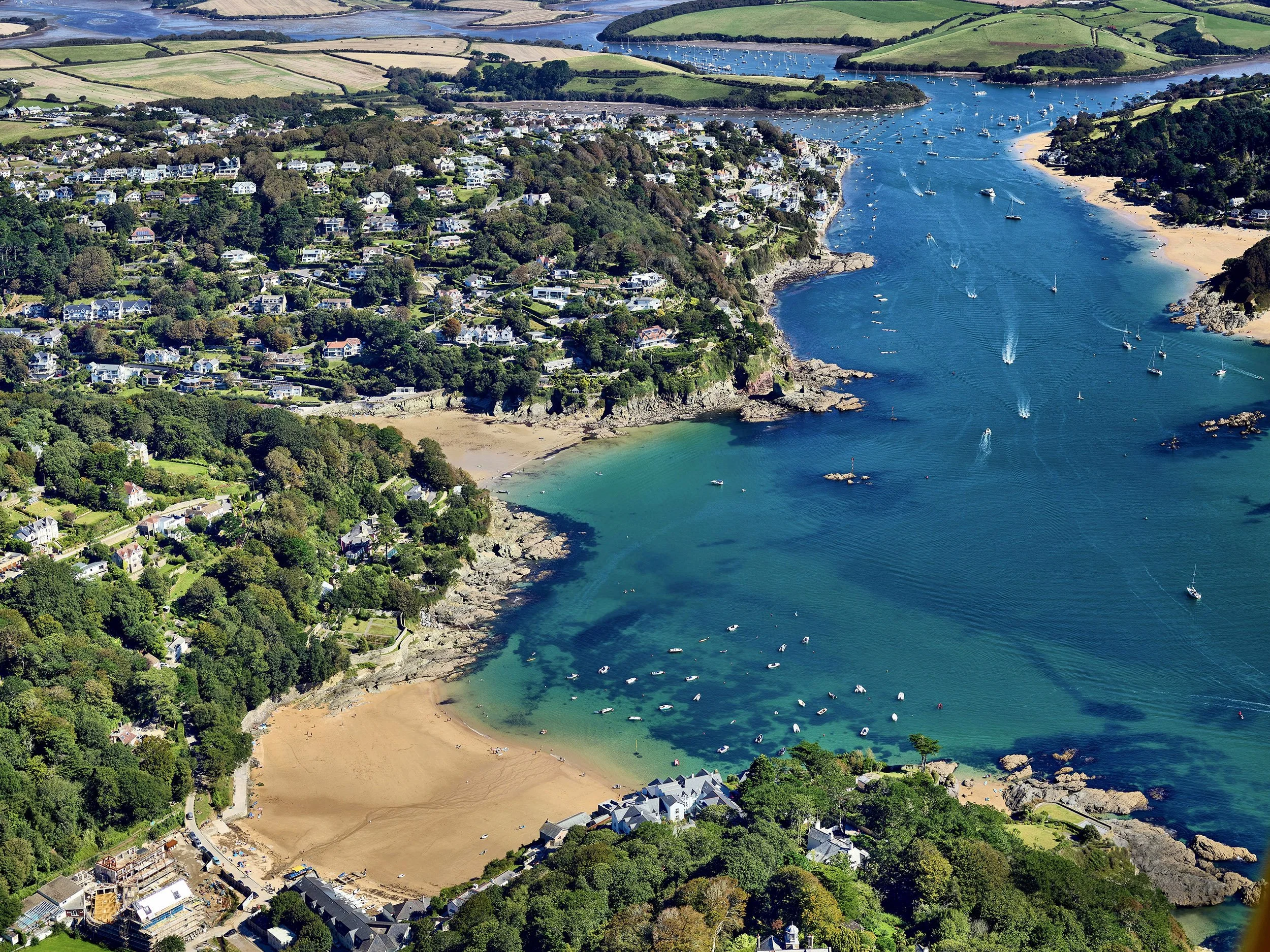 Aerial view of a coastal town with a sandy beach, numerous boats in the water, green hills, and residential homes surrounded by trees.