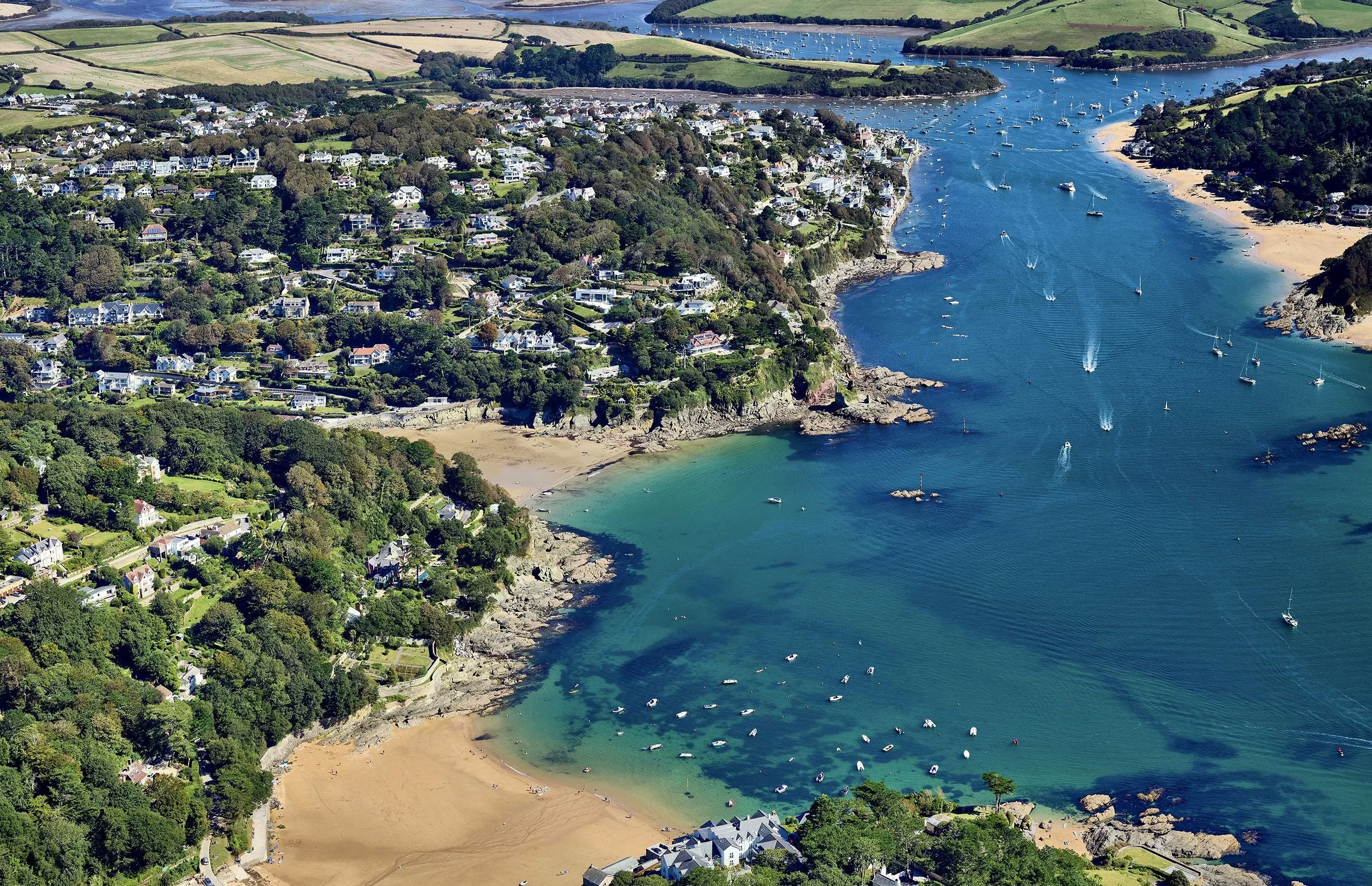 Aerial view of a coastal area with a beach, boats in the water, and a residential neighborhood surrounded by greenery.