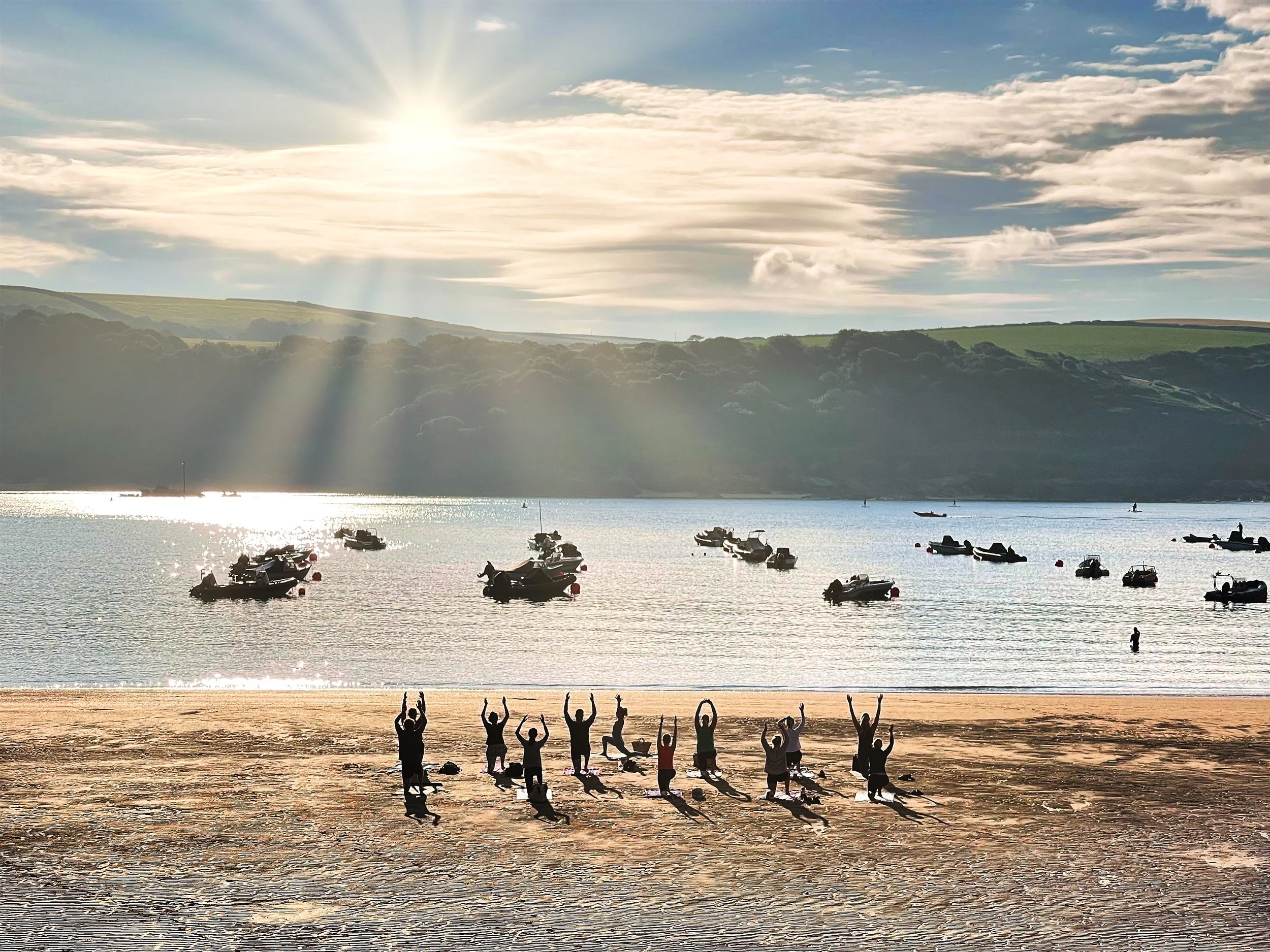 Group of people practicing yoga on a sandy beach near the water, with boats floating on calm water and hills in the background, under a bright sun with some clouds.
