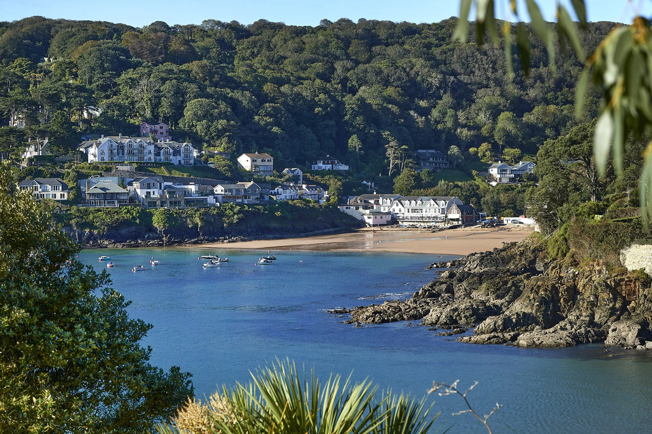 Coastal view of a bay with boats anchored in the water, a sandy beach, rocky shoreline, and residential buildings on a hillside surrounded by lush greenery.