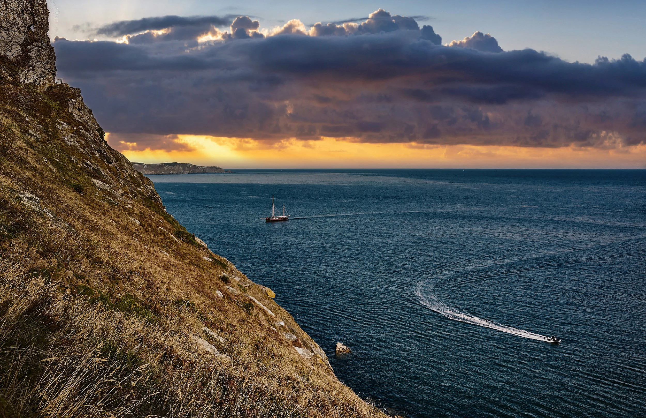 A coastal scene at sunset with a steep grassy and rocky cliff on the left, a sailing boat and a motorboat in the water, and dark clouds with a break in the sky revealing a warm glow over the horizon.