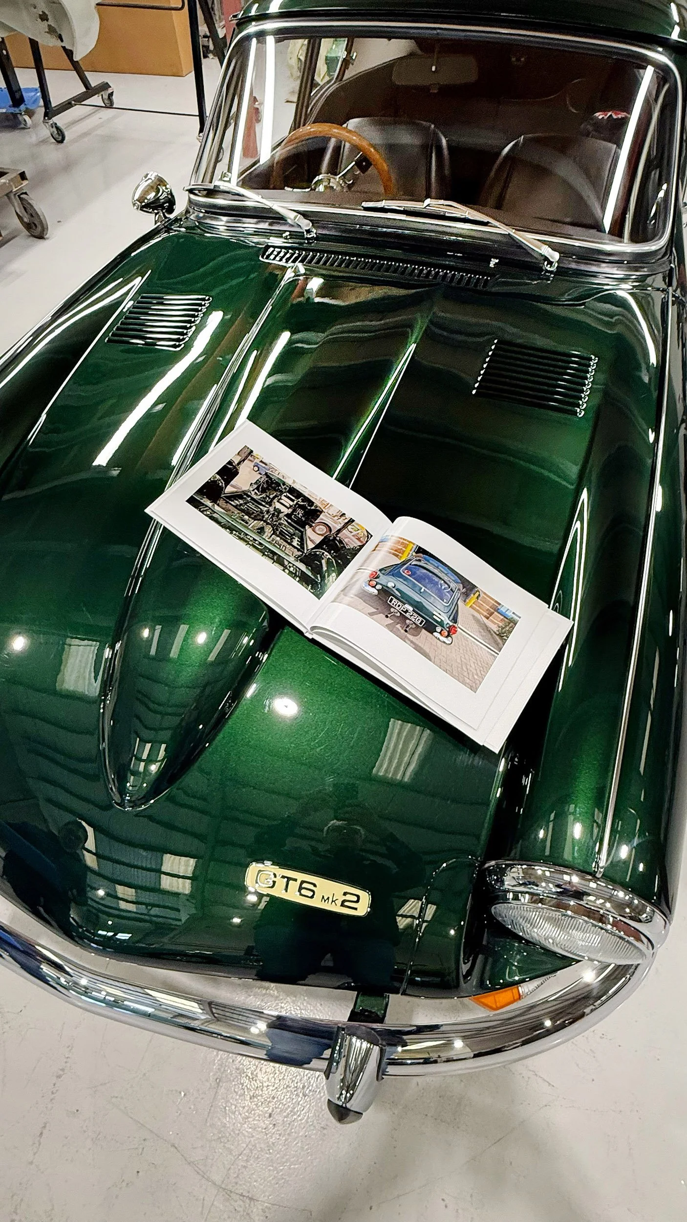 A classic green vintage sports car with a wooden steering wheel, displayed indoors on a shiny floor. An open book with photographs of the car's engine and rear view is placed on the hood.