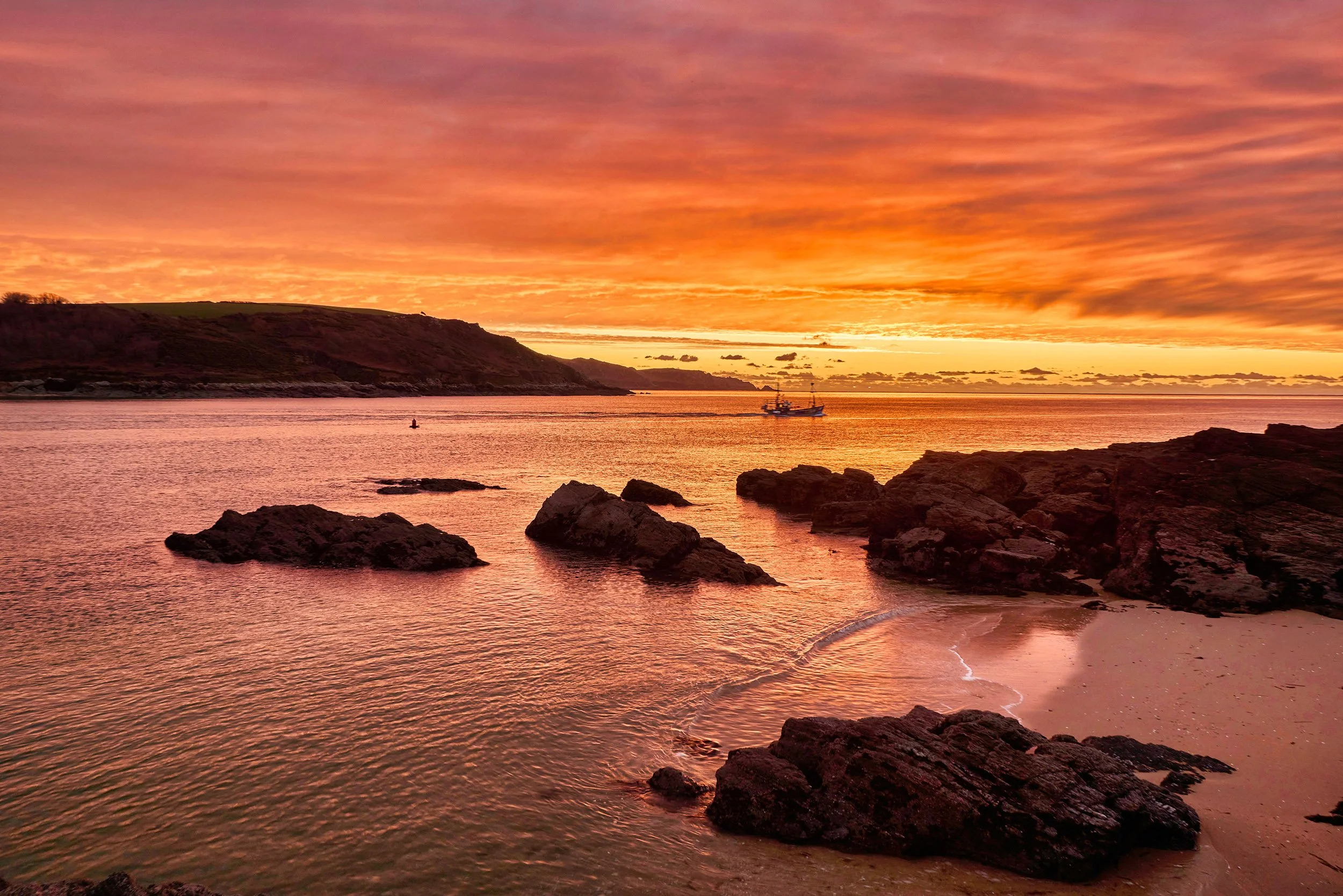 A sunset over a rocky beach with a boat on the water and a colorful sky.