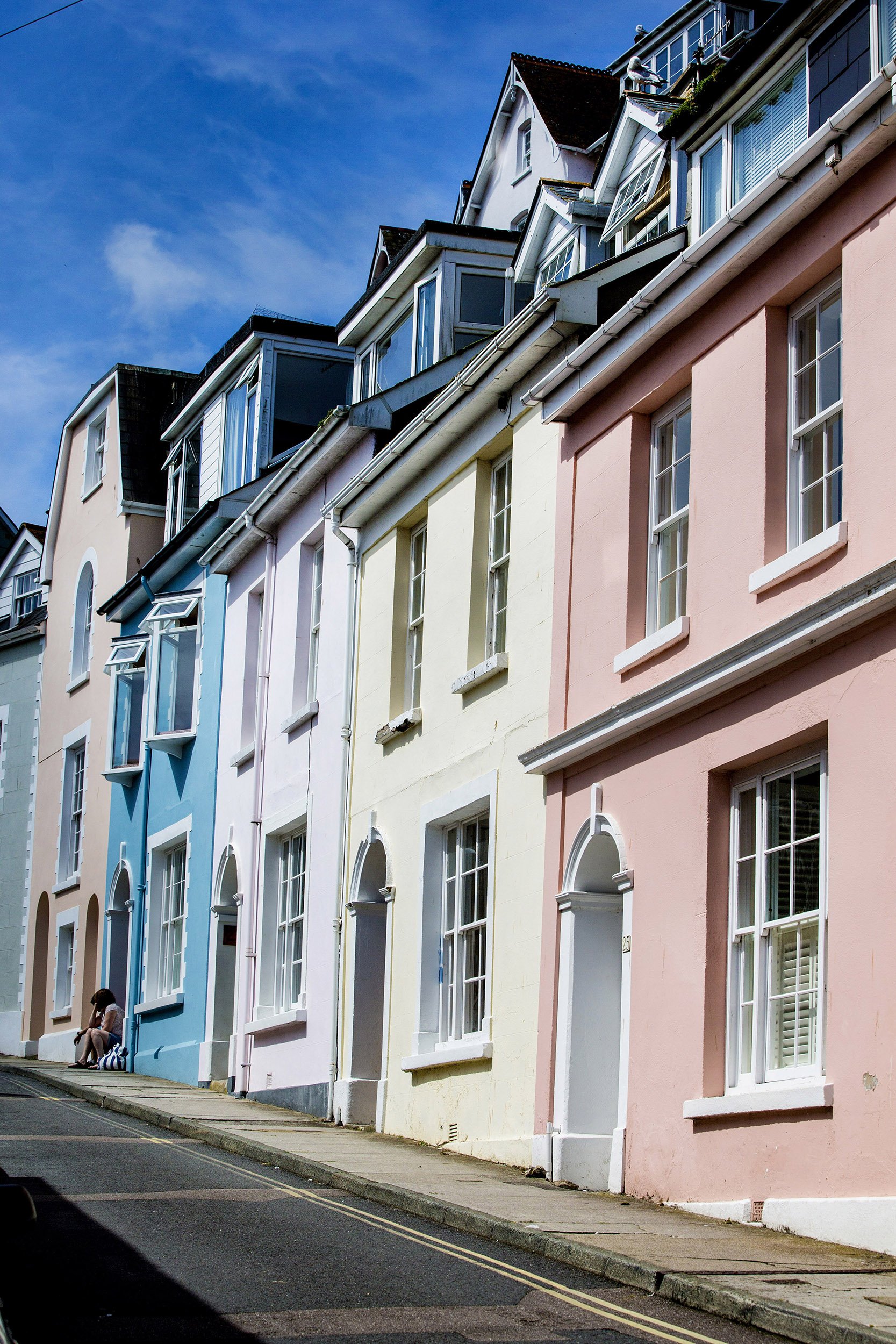 Colorful pastel row houses with unique architecture on a sidewalk under a bright blue sky, with a person sitting on the curb.