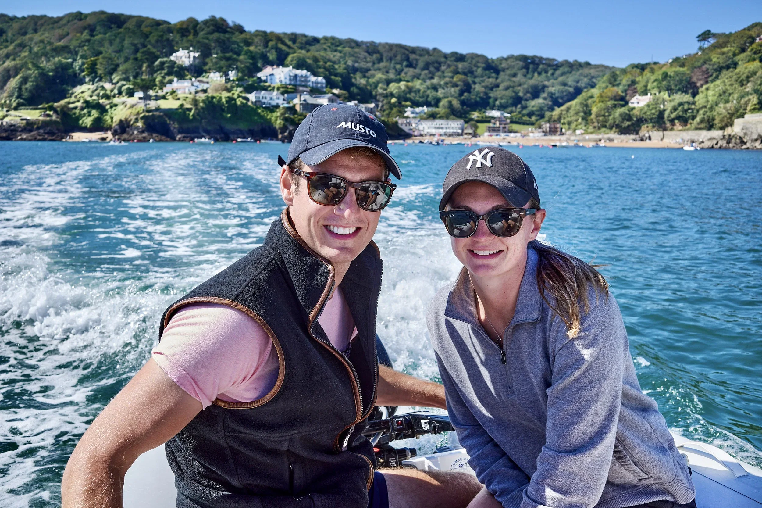 Two smiling people wearing sunglasses and hats, riding on a boat on the water with a shoreline and houses in the background.