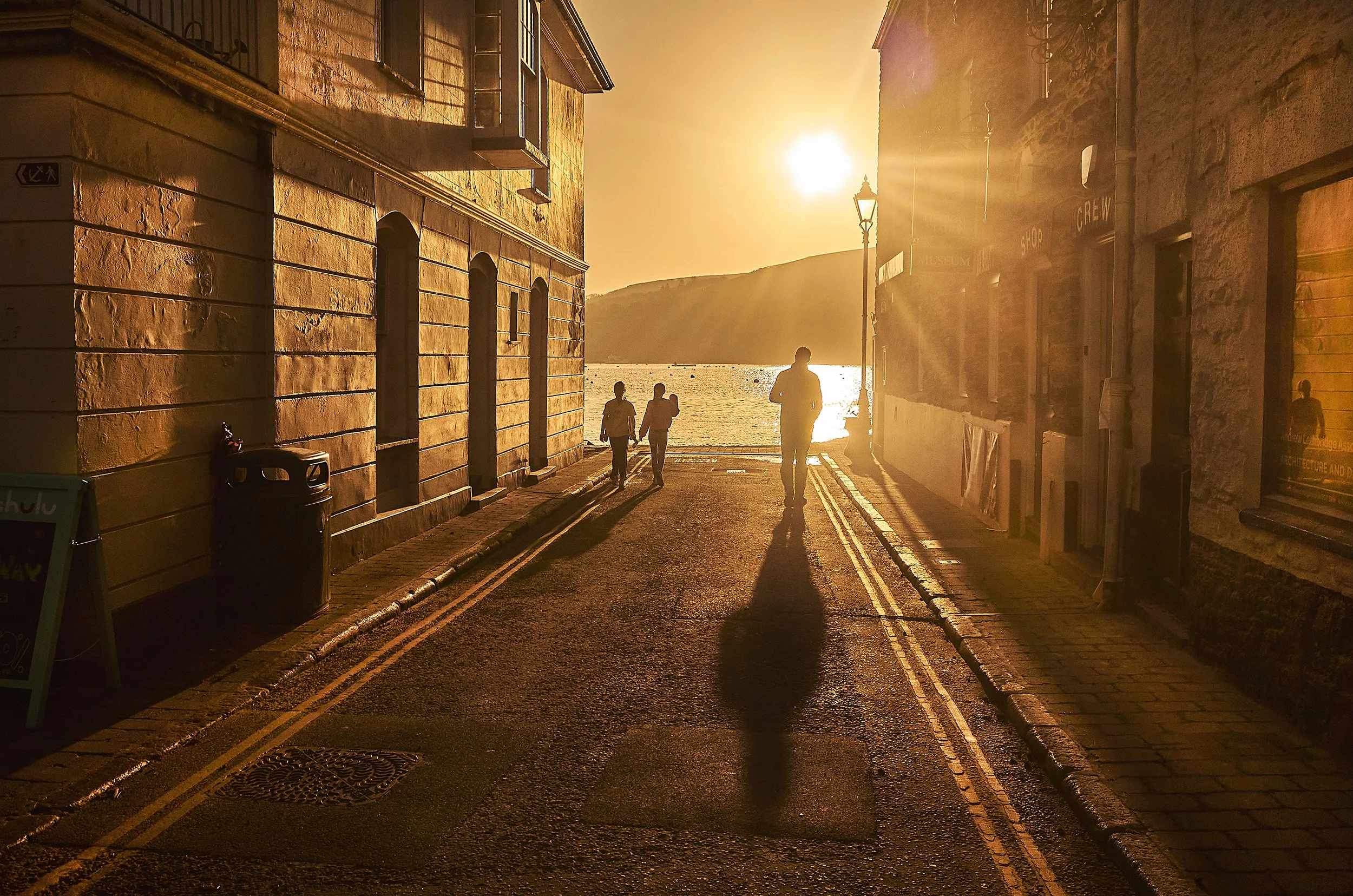 Silhouetted people walking on a street near a body of water during sunset, with buildings on either side and hills in the background.