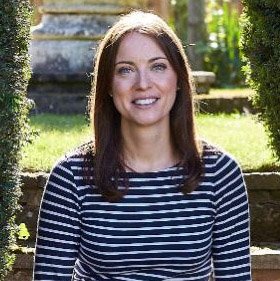 A young woman with long brown hair, smiling, wearing a navy and white striped shirt, standing outdoors with greenery and stone steps in the background.