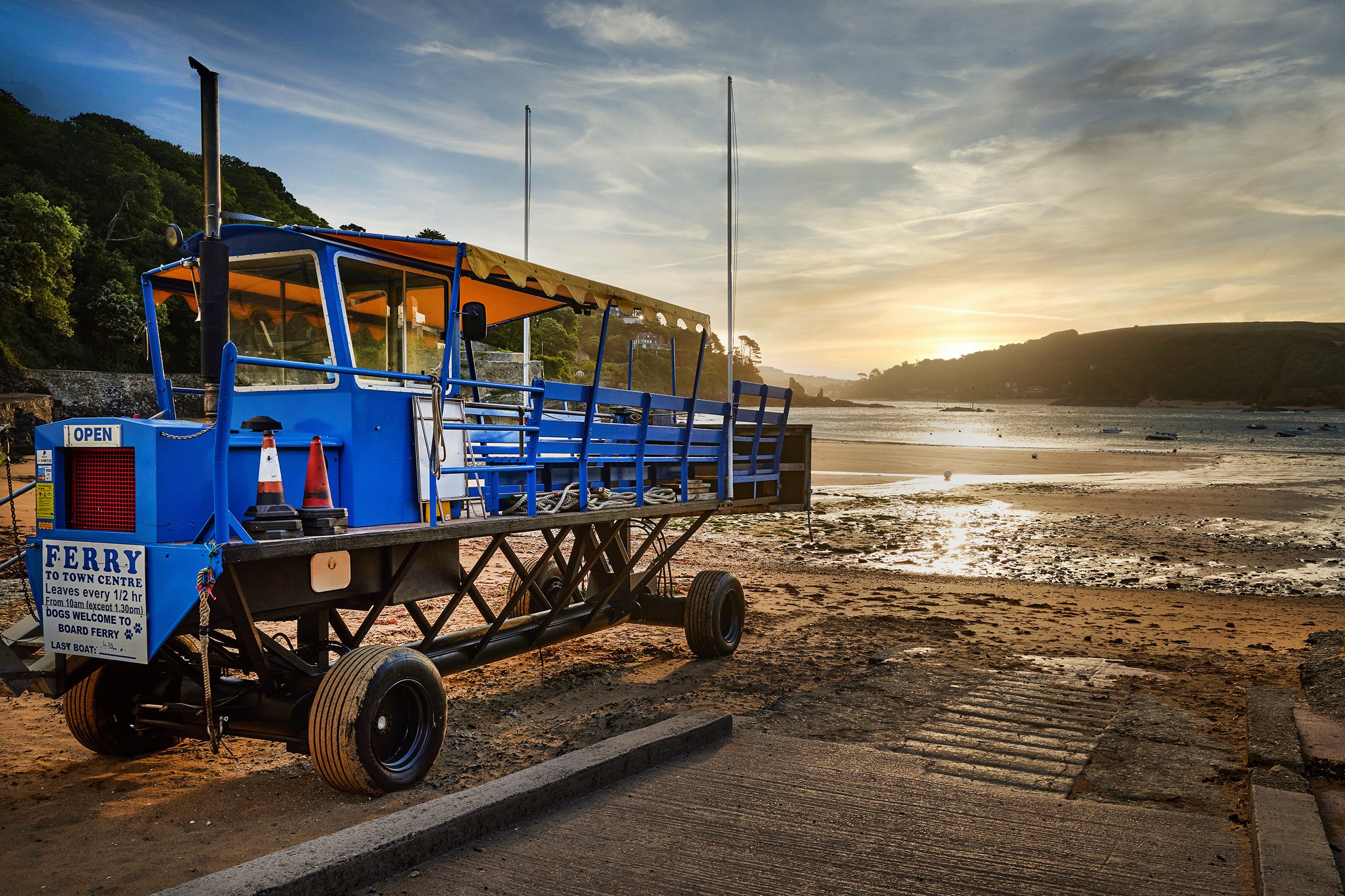 Blue ferry boat on the shore at sunset with hills and water in the background.