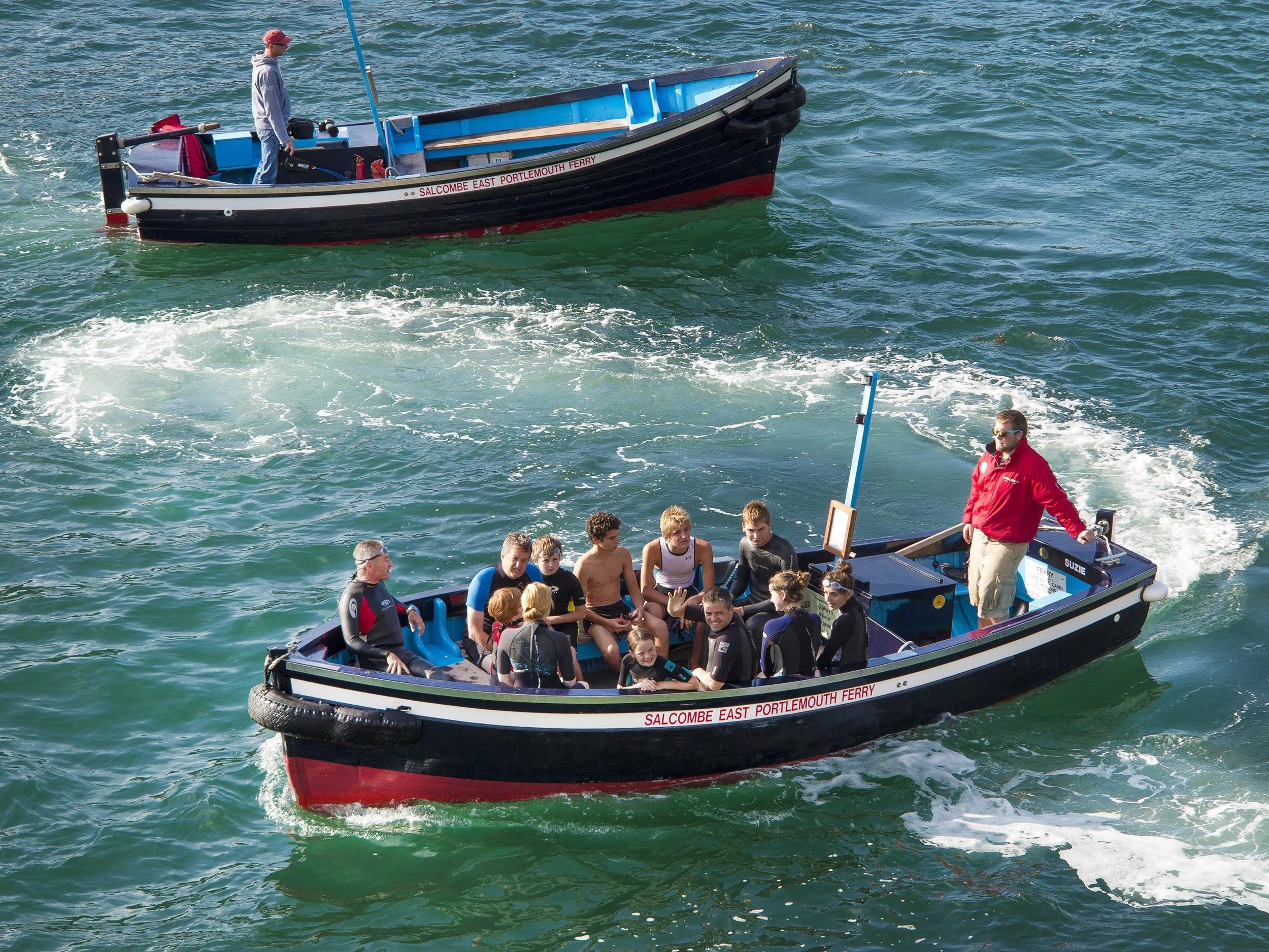 Two boats on the water, one with a group of people seated, and the other with a person at the helm. The boats are part of the Salcombe East Portlemouth Ferry service.