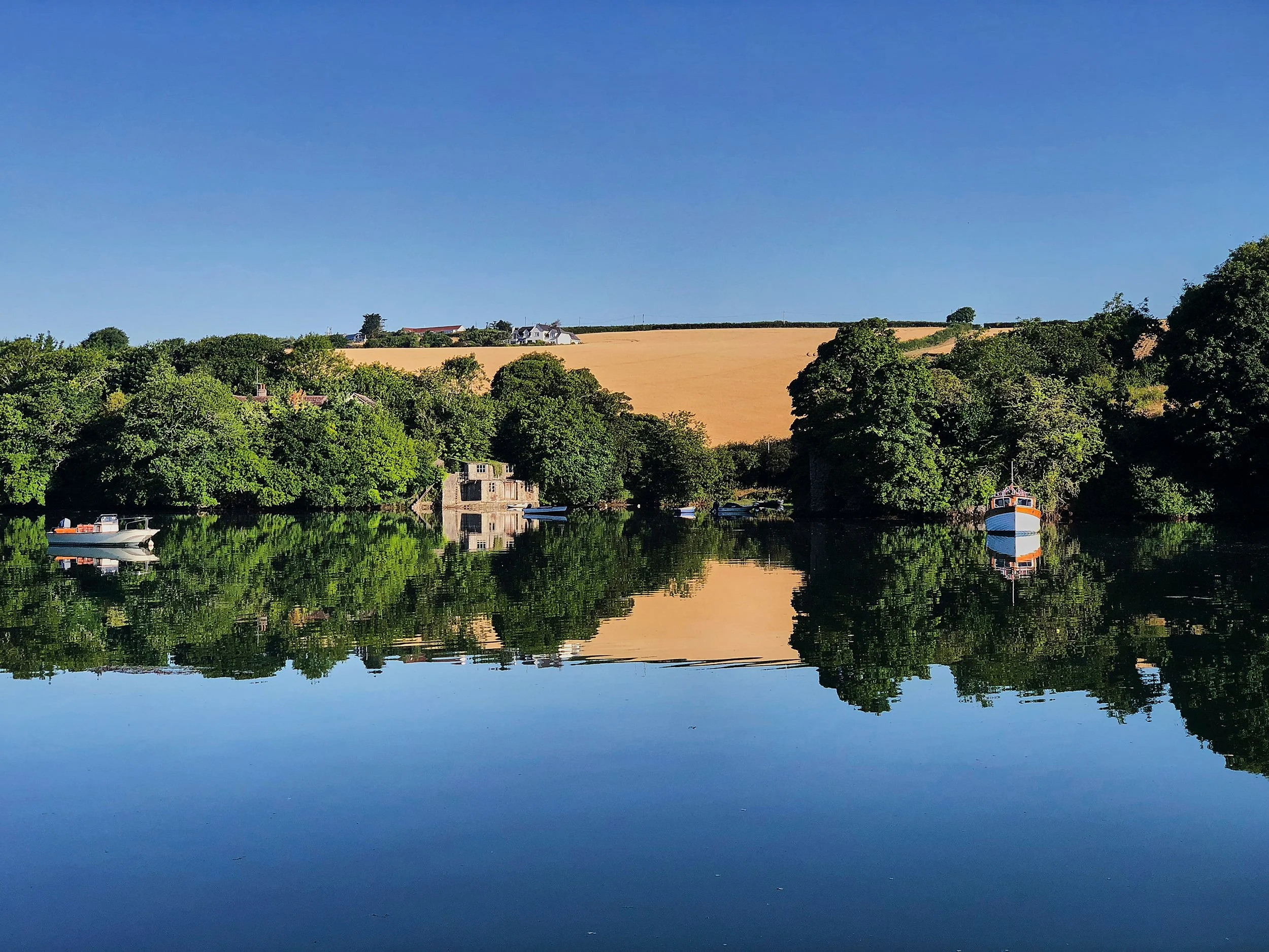 Calm river reflecting green trees, boats, and a building, with rolling hills and houses in the background under a clear blue sky.