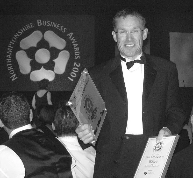 A man in a tuxedo holding two awards at the 2011 North Hampshire Business Awards. There are other people seated at tables in the background.