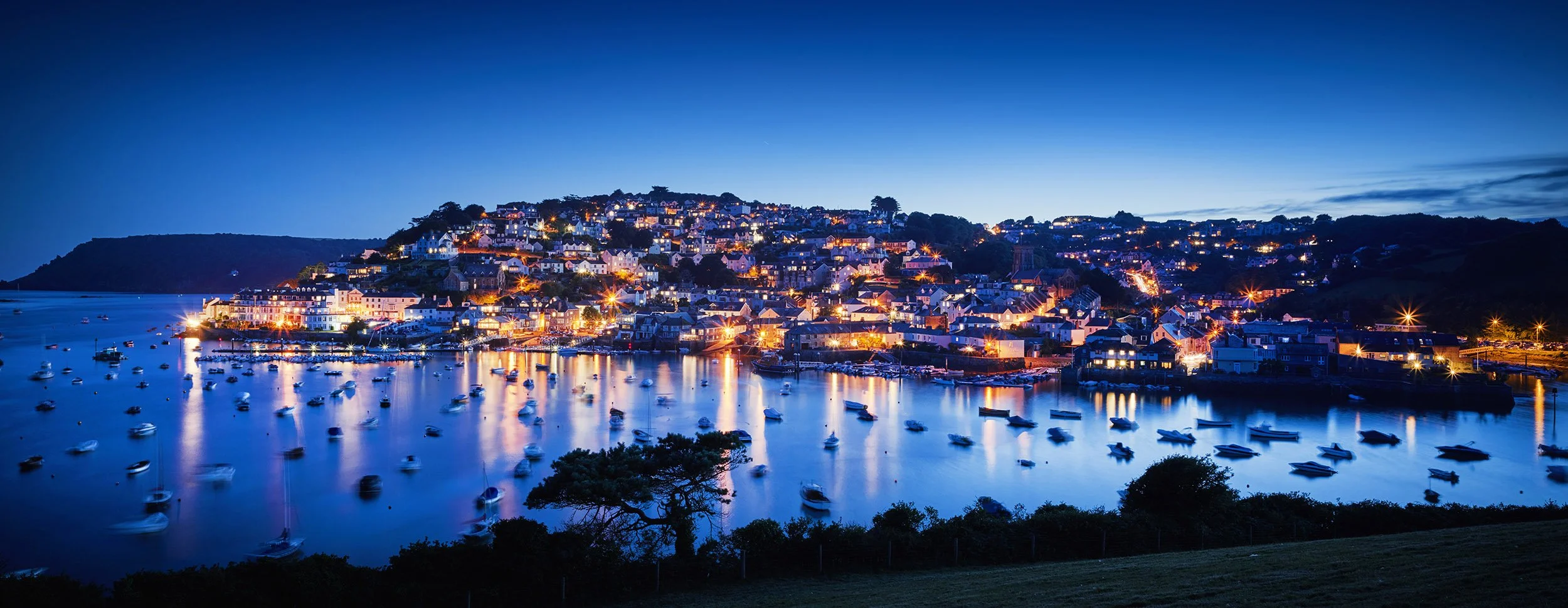 A coastal town during dusk with numerous boats anchored in the calm water, illuminated buildings on the hill, and a clear blue sky.