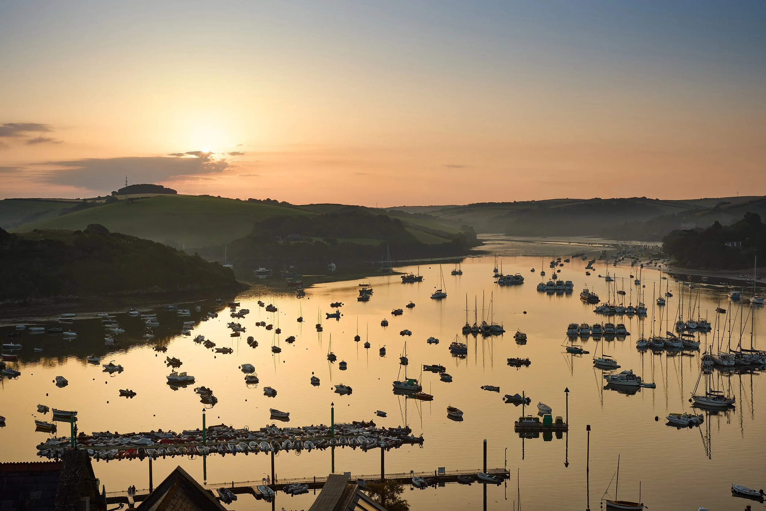 Sunset over a harbor with numerous boats anchored in calm water, green hills in the background, and a partly cloudy sky.